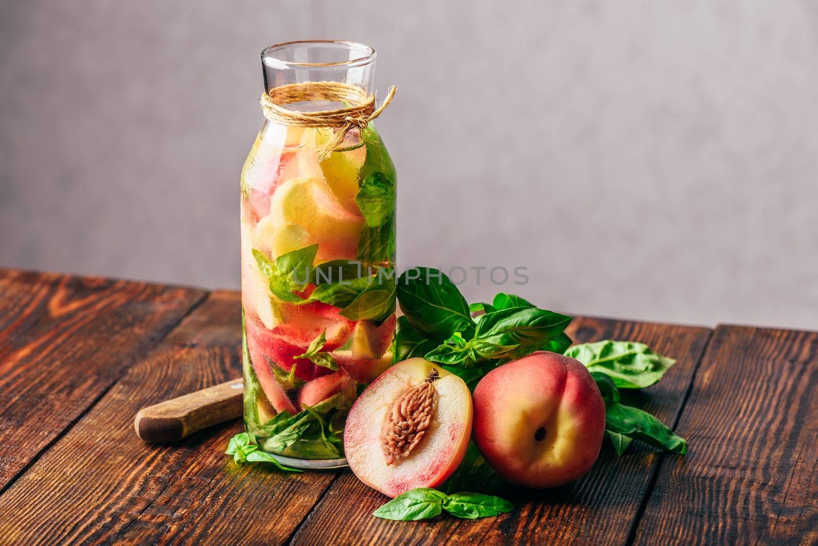 Bottle of Summer Water with Sliced Peach and Basil Leaves. Knife and Ingredients on Wooden Table.