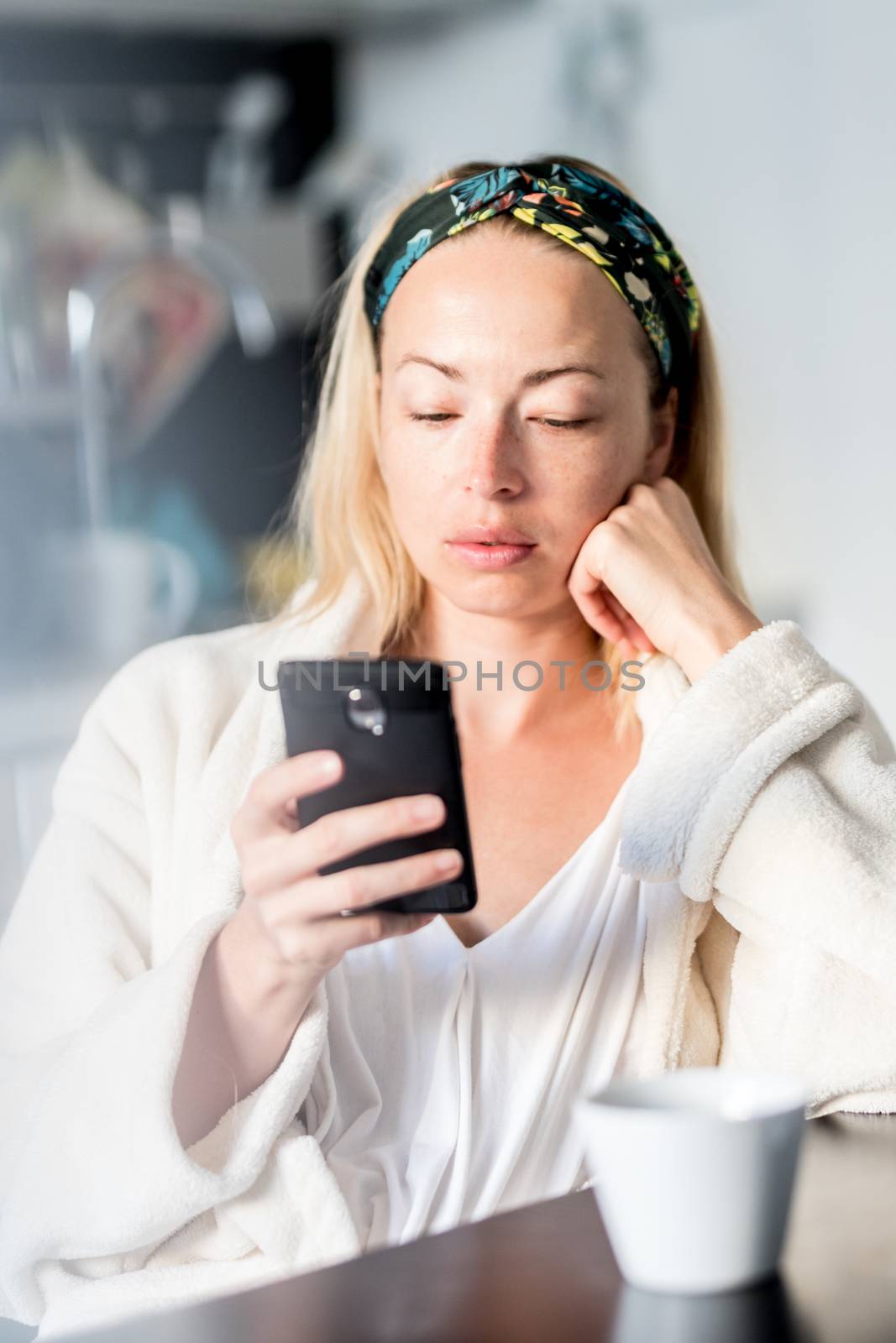 Beautiful caucasian woman at home, feeling comfortable wearing white bathrobe, taking some time to herself, drinking morning coffee and reading news on mobile phone device in the morning by kasto