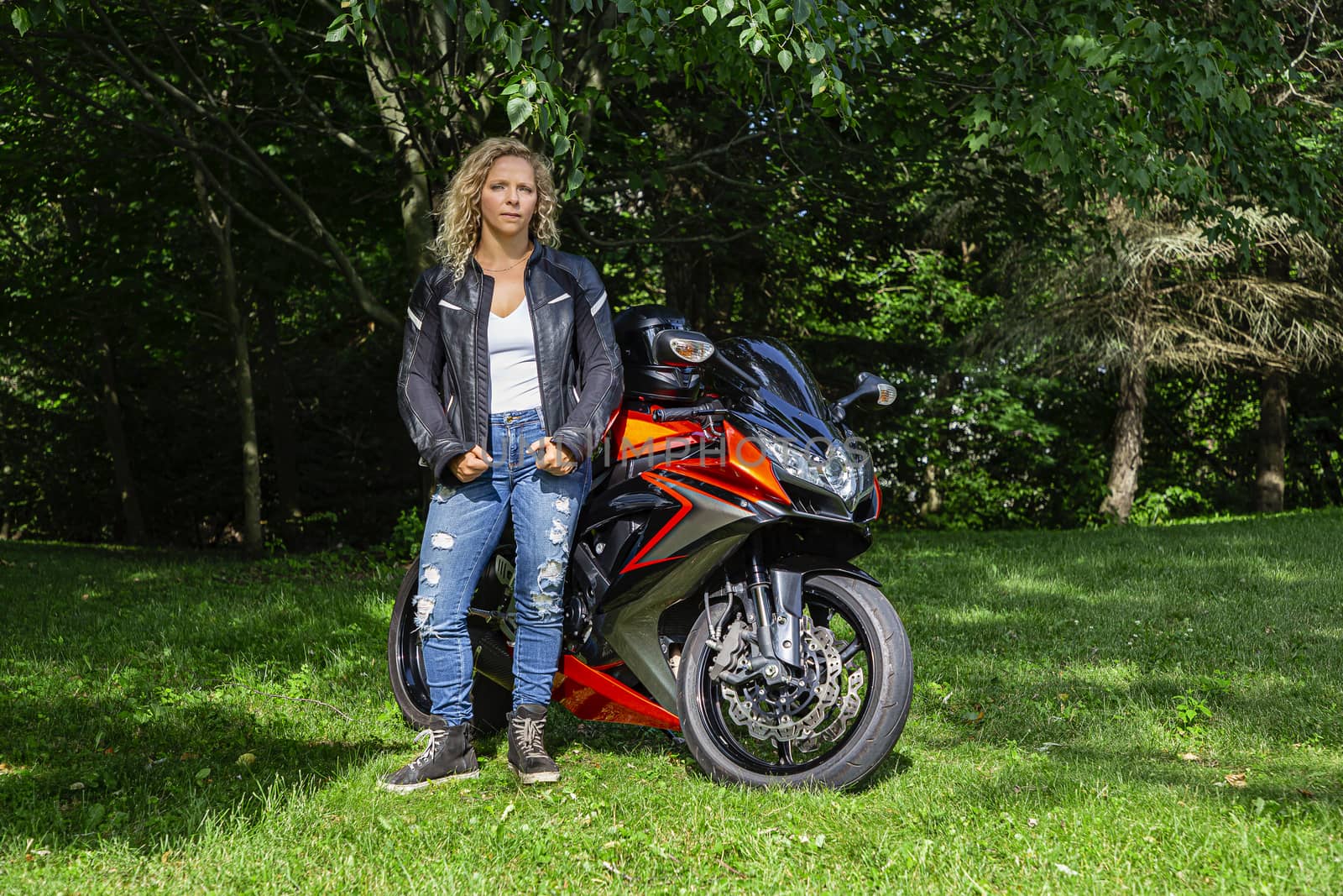 young blond woman, wearing a black leather jacket, rip jeans and tank top, standing in front of a motocycle, in nature