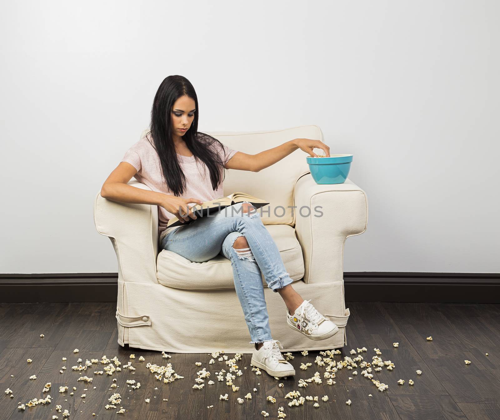 young woman, sitting on a white couch, with popcorn all over the floor, reading a book and eating popcorn