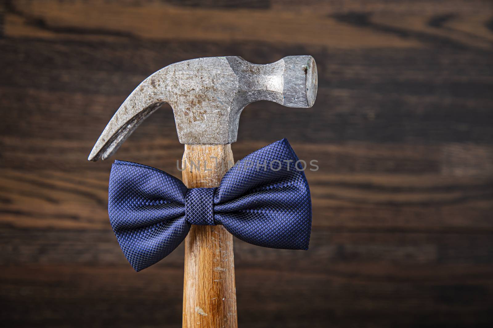 Old hammer with wood handle, wearing a blue bow tie, against a dark wood background