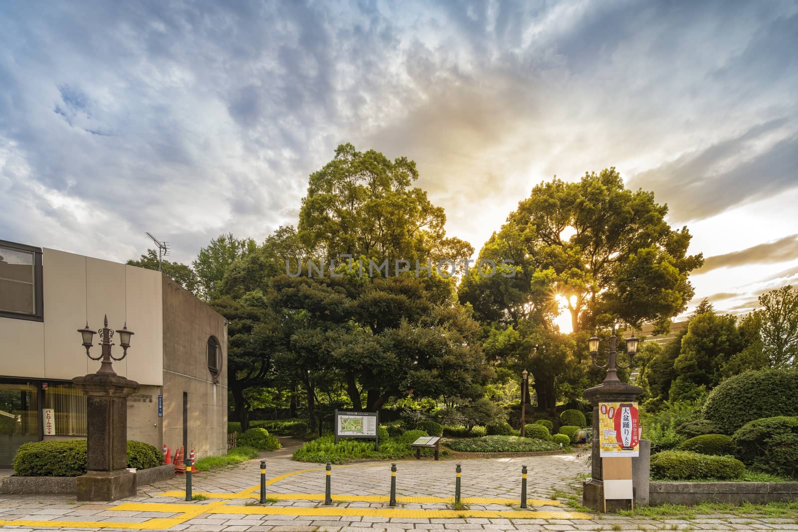 Sunset sky on the entrance of the Hibiya Park (日比谷公園 Hibiya Kōen) in Chiyoda City of Tokyo in Japan. Its entrance is decorated with Gothic cast iron lanterns from the 1930s. The poster on the right pillar informs about the Obon Festival that takes place every year in summer to celebrate the deceased ancestors. The modern building on the left is a police station where a sign indicates in red characters that the agent is currently on patrol.