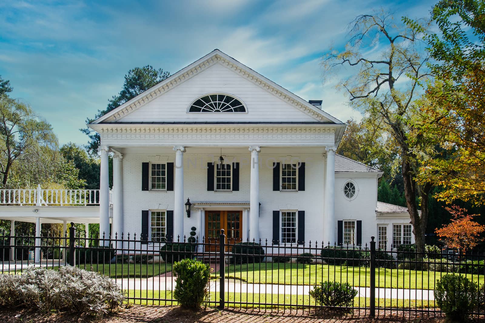 A traditional white home with columns and carport in a landscaped lawn behind a black wrought iron fence