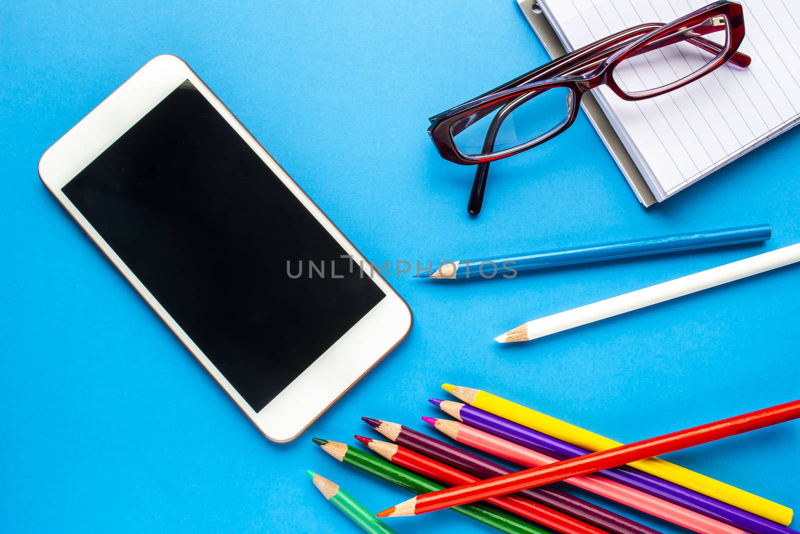 A smartphone on a blue background with colour pencils, notebook and glasses