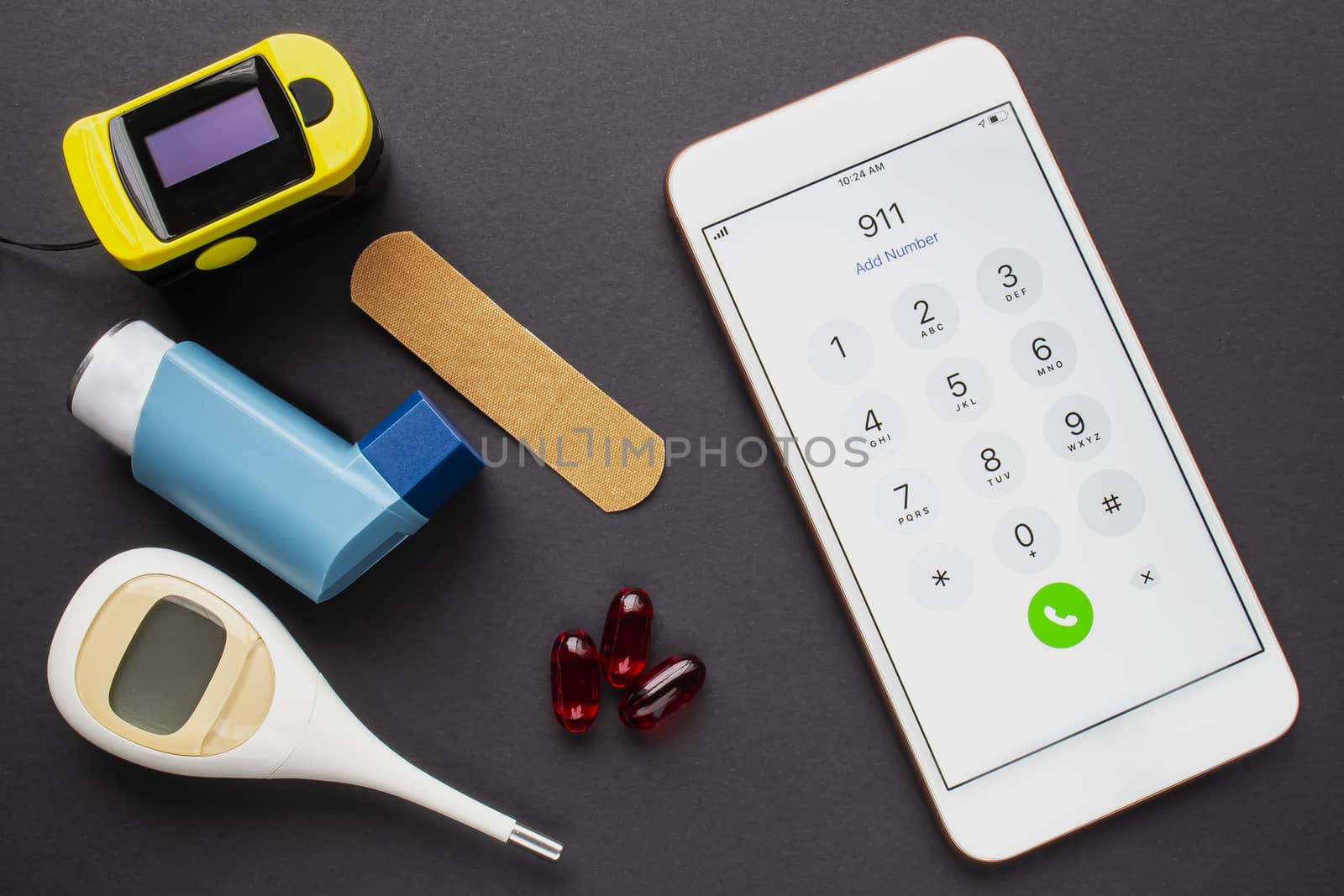 An smartphone calling the 911 dial emergency services, with an asthma inhaler, a bandage, some pills, and an oximeter on a black background.