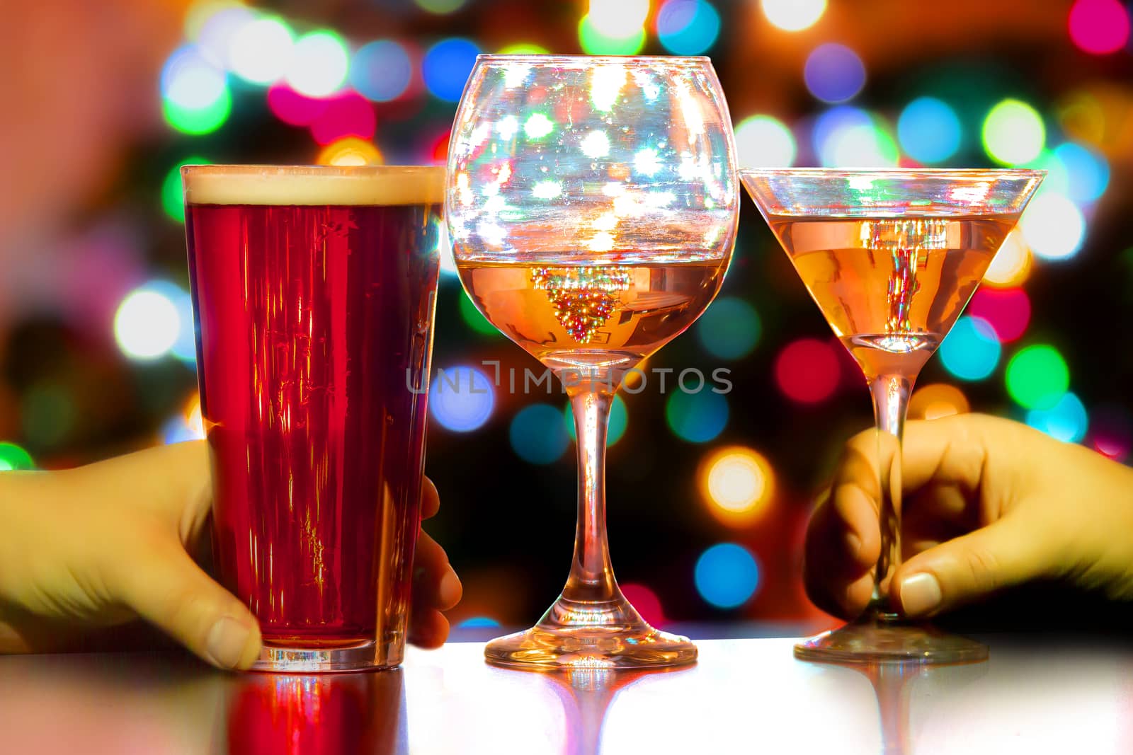 A close up of a couple of people's hand with a beer, wine and a martini and colourful lights on the background