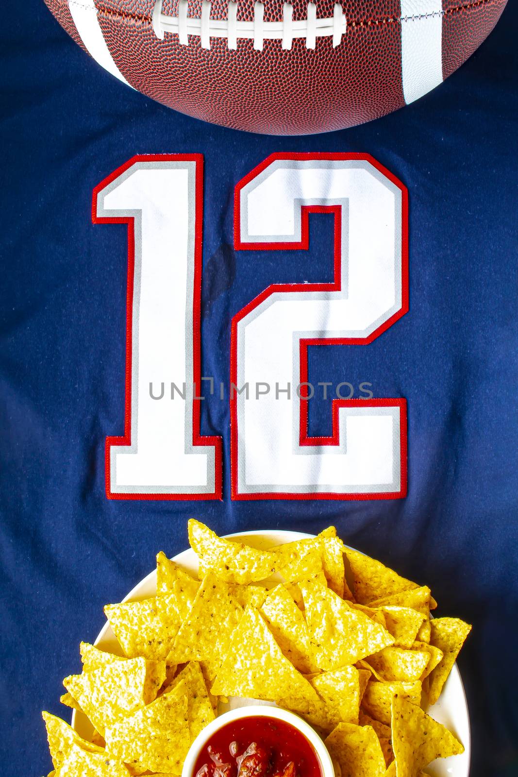 An American football with organic nacho chips and mild salsa on a white blue football jersey with the 12 number on vertical view by oasisamuel