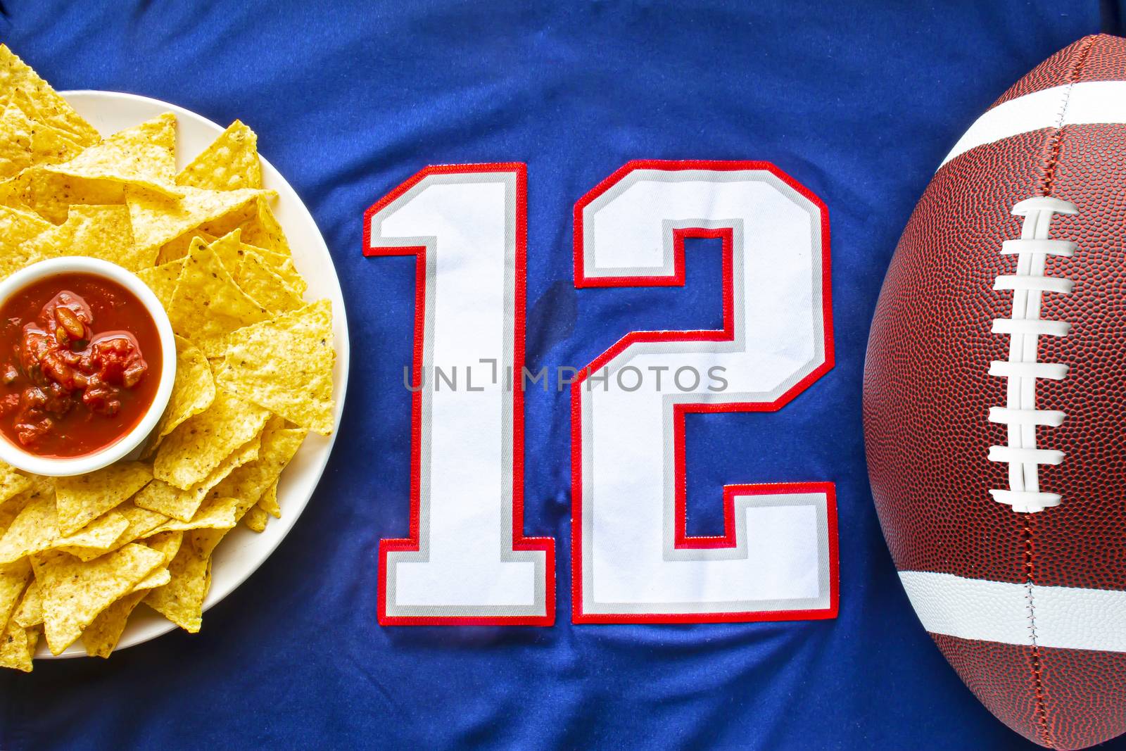 An American football with organic nacho chips and mild salsa on a white blue football jersey with the 12 number on horizontal view