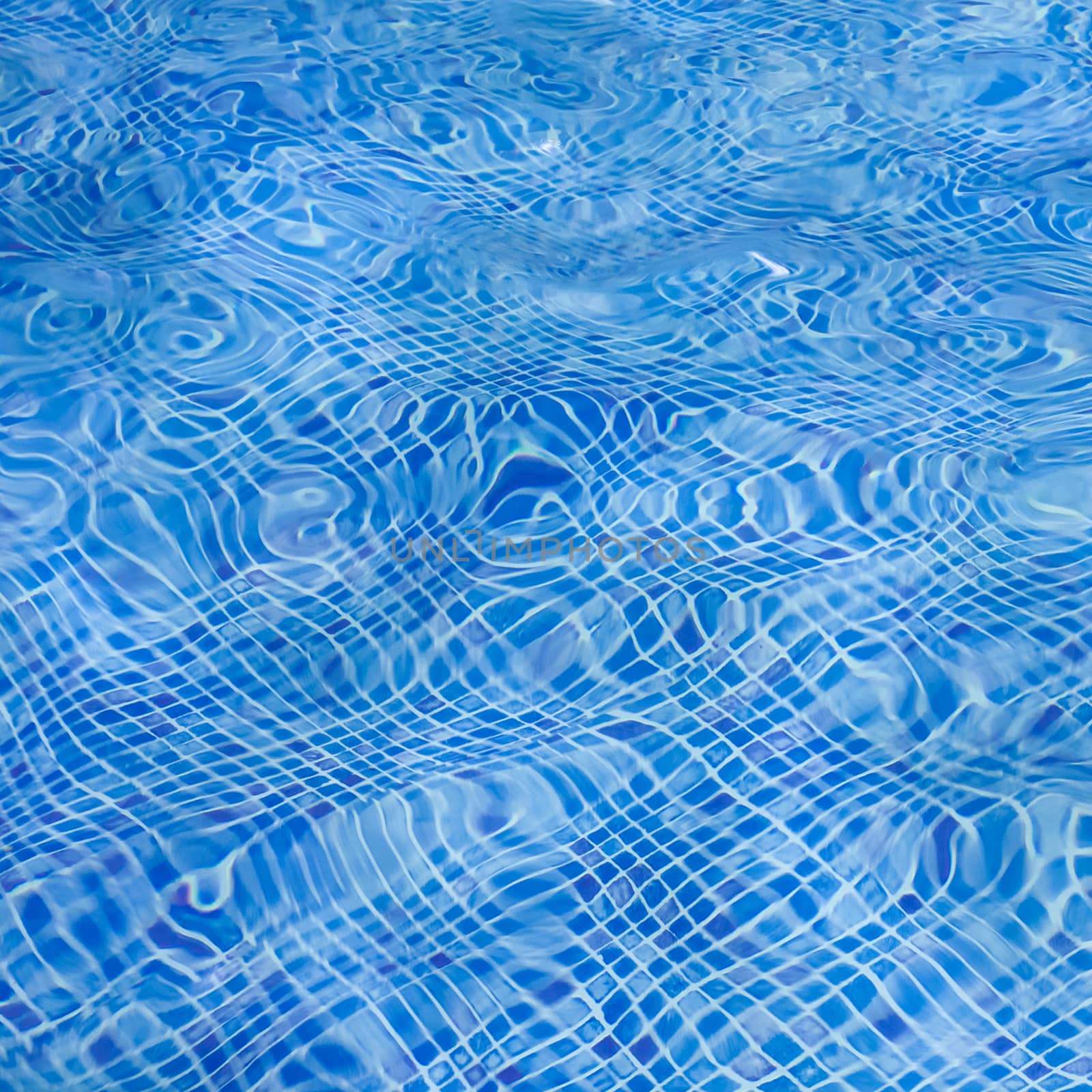 The light reflected on the top of the water surface of the pool lined with blue mosaic tiles below. Water ripples on blue-tiled swimming pool background.