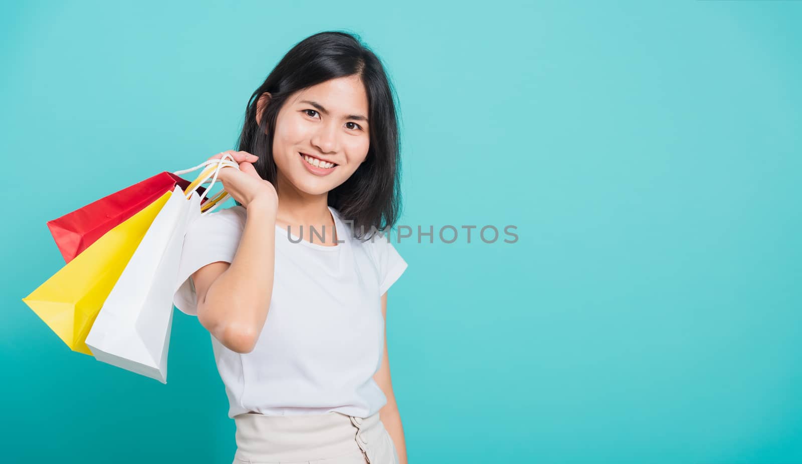 Portrait happy Asian beautiful young woman standing wear white t-shirt, She holding shopping bags multi color on hand and looking to camera, shoot photo in a studio on a blue background