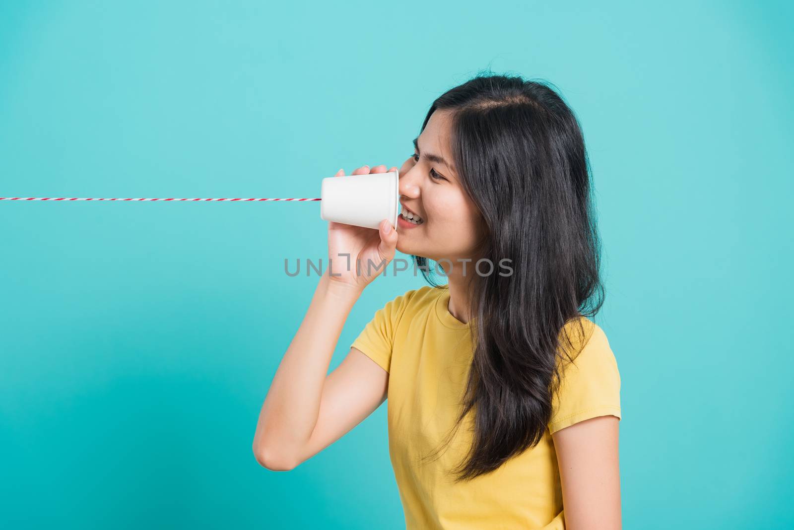 Portrait happy Asian beautiful young woman smile white teeth standing wear yellow t-shirt, She holding paper can telephone for talking, studio shot on blue background with copy space for text