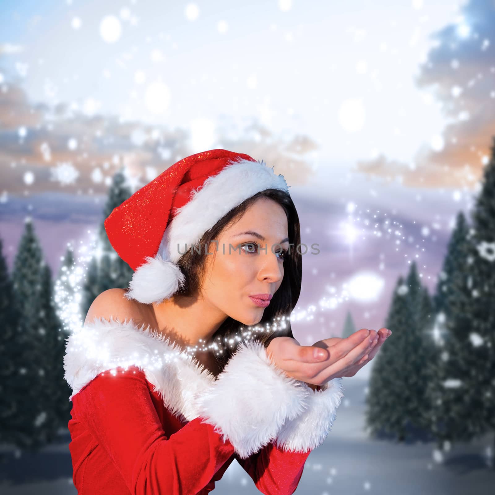 Pretty girl in santa outfit blowing against snowy landscape with fir trees