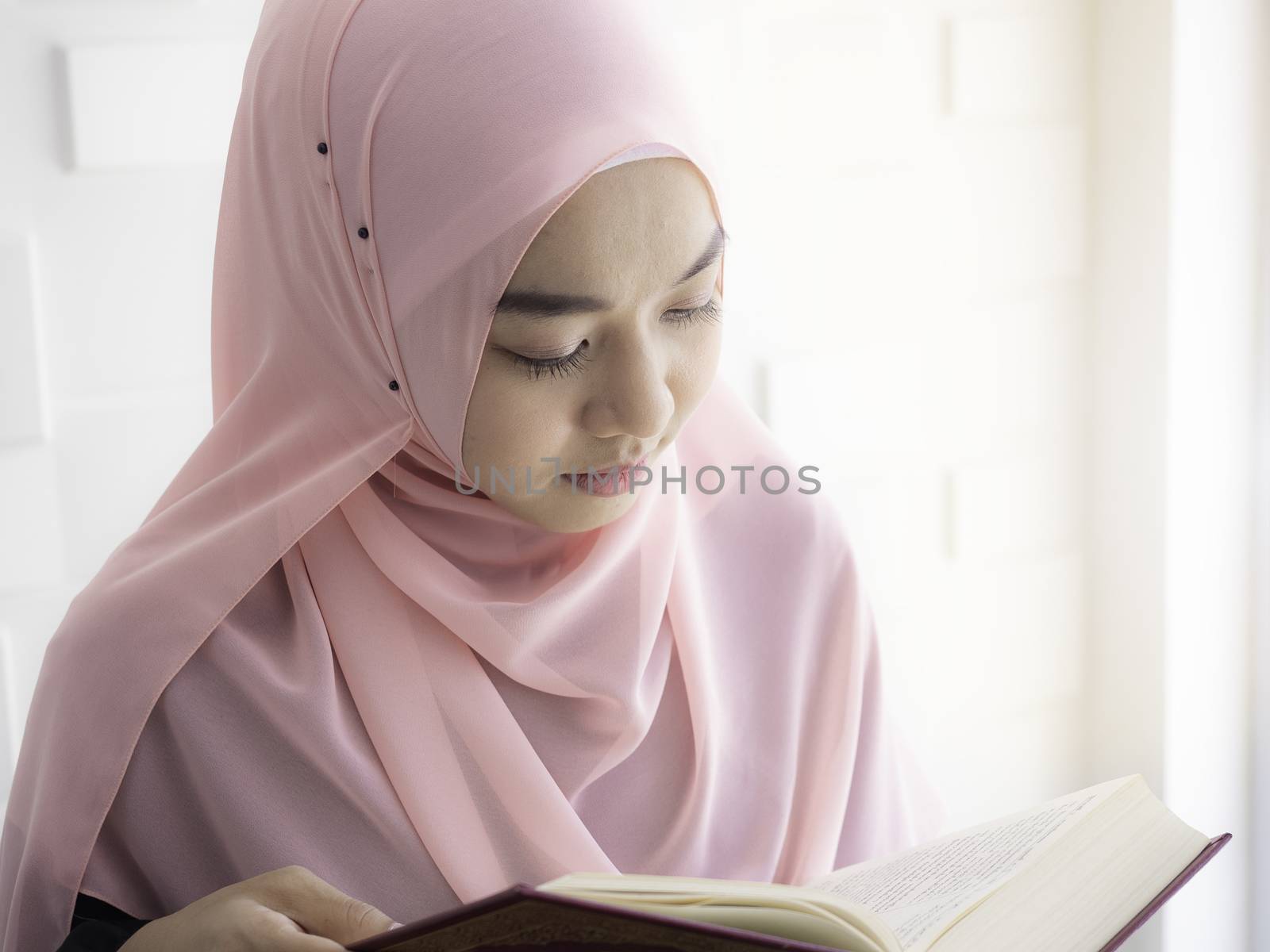 An Asian Muslim woman is concentrating on reading the Al Quran, concentrating carefully in the mosque with the warm sunlight shining through the windows.