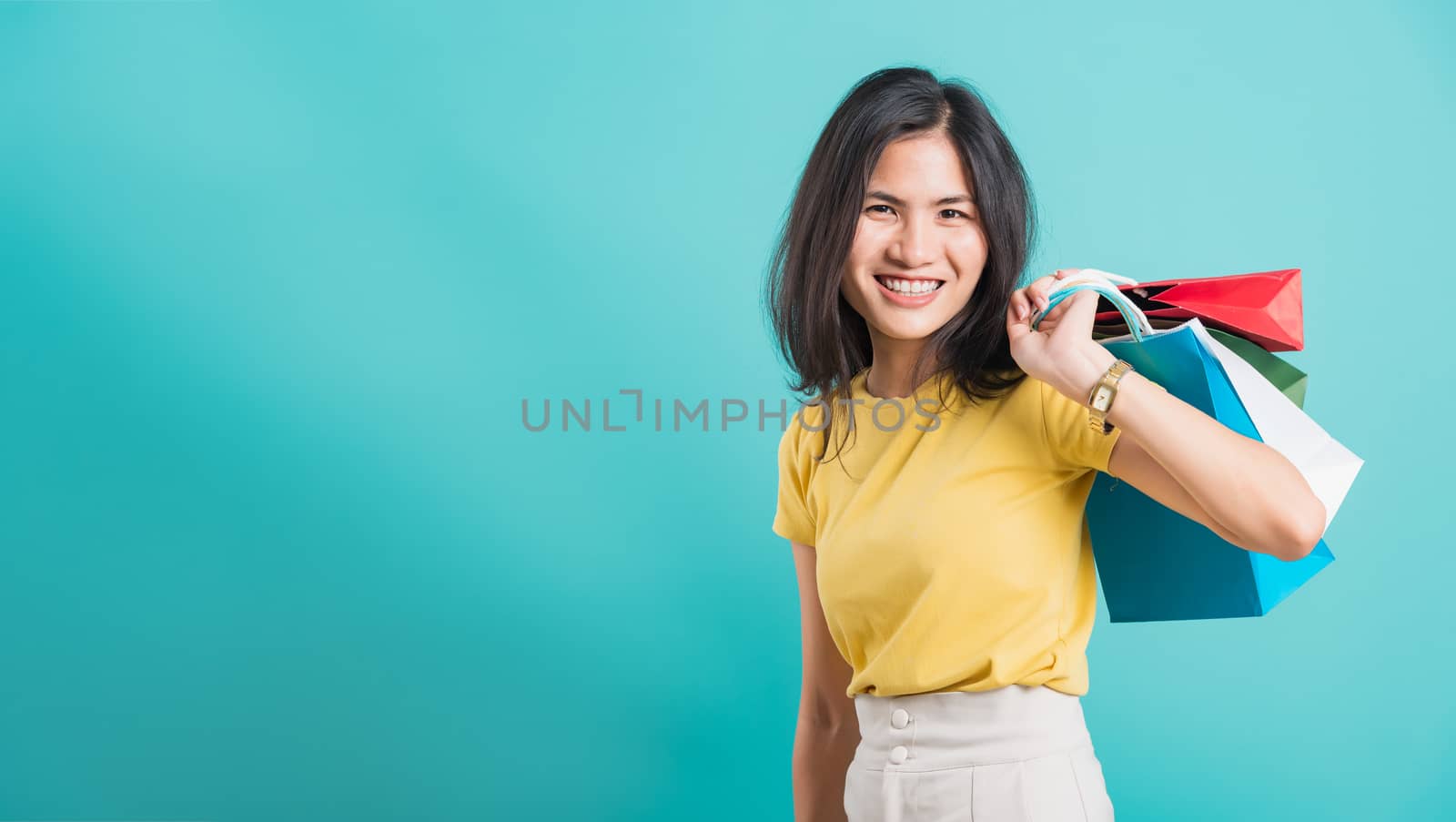 Portrait happy Asian beautiful young woman smile white teeth standing wear yellow t-shirt, She holding shopping bags on hand her looking camera, shoot photo in studio on blue background with copyspace