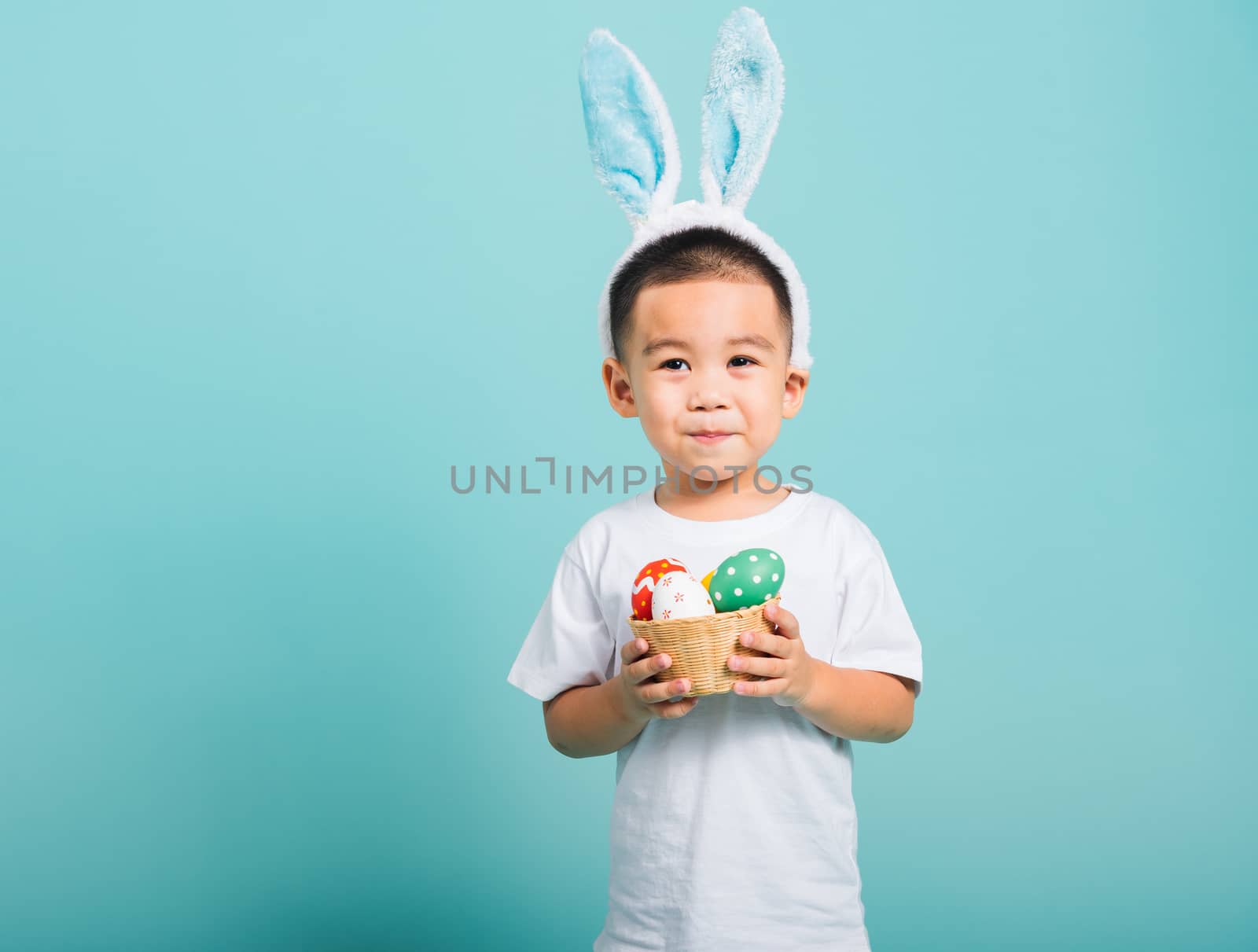 Asian cute little child boy smile beaming wearing bunny ears and a white T-shirt, standing to hold a basket with full Easter eggs on blue background with copy space