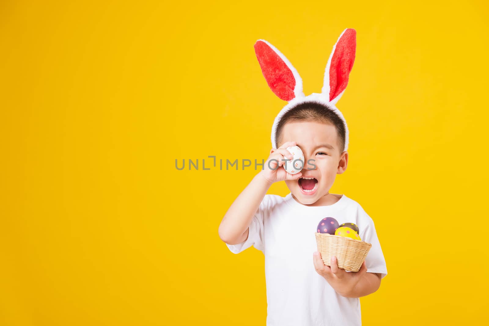 Portrait happy Asian cute little children boy smile standing so happy wearing white T-shirt and bunny ears in Easter festival day holding easter eggs, studio shot on yellow background with copy space