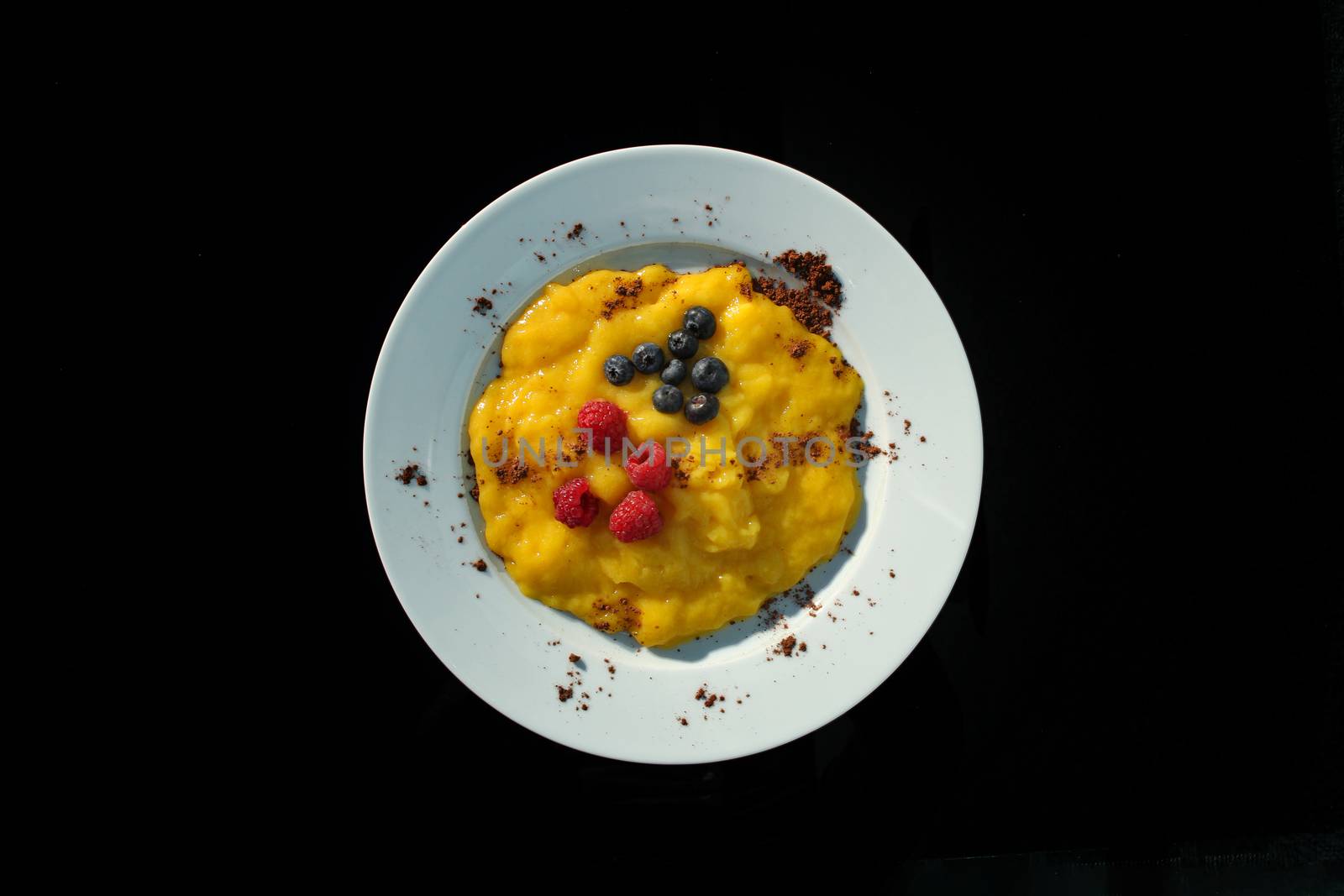 Yellow pudding with blueberry and raspberries with chocolate chips on a white plate and black background