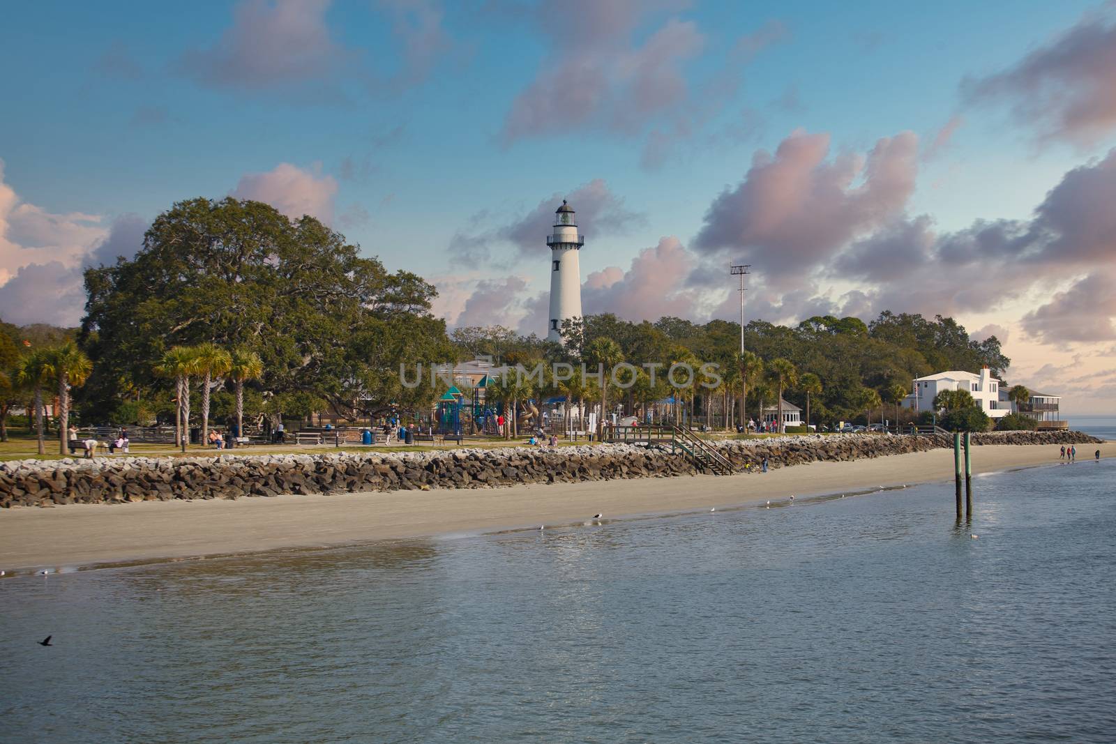 A white brick lighthouse beyond a nice park full of people enjoying the beach