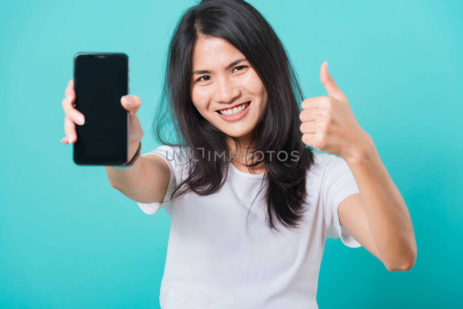 Portrait Asian beautiful happy young woman standing smile, holding blank screen mobile phone and showing thumbs up gesture, shoot photo in studio focus face on blue background, with copy space