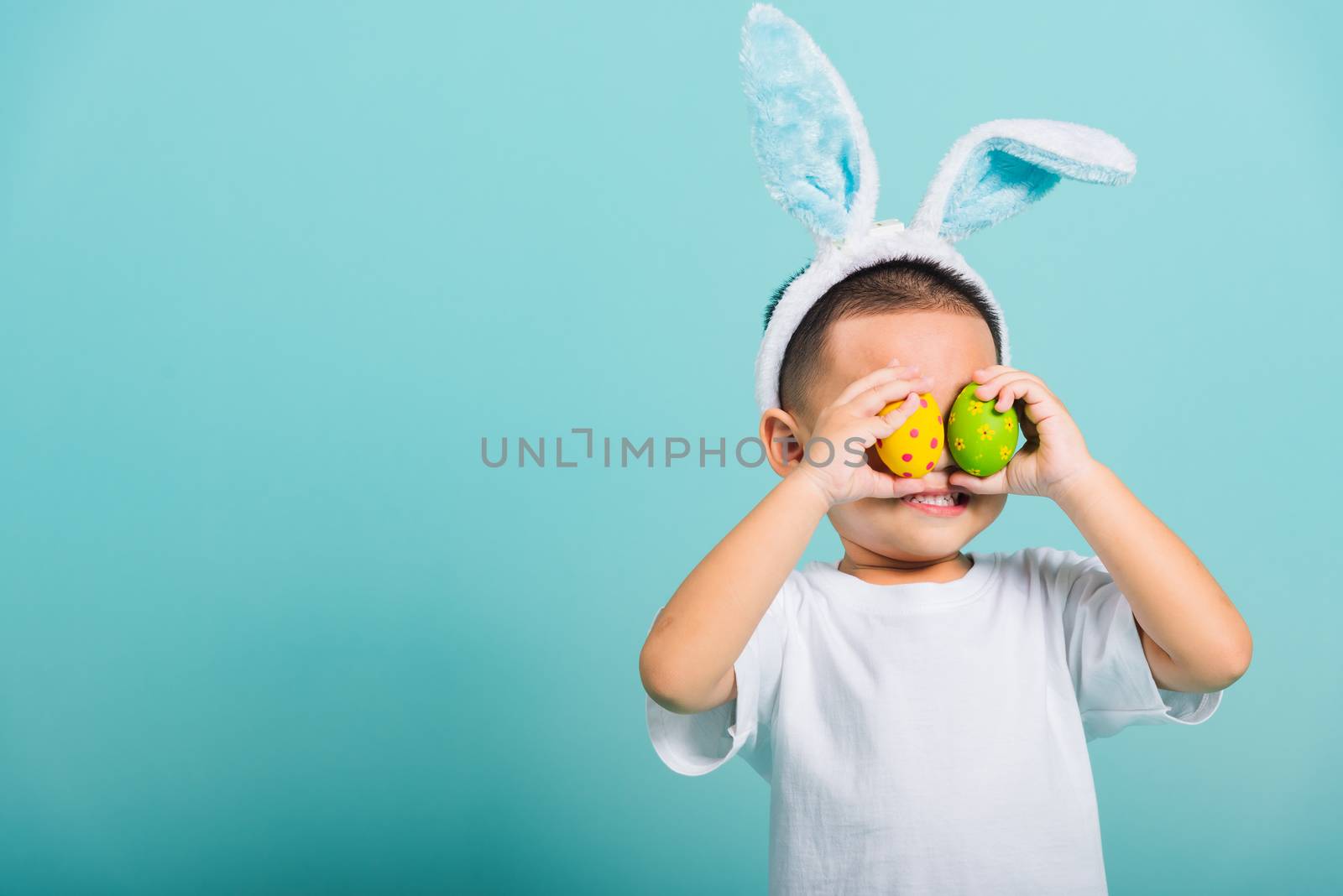 Asian cute little child boy smile beaming wearing bunny ears and a white T-shirt, standing to holds colored easter eggs instead of eyes on blue background with copy space for text