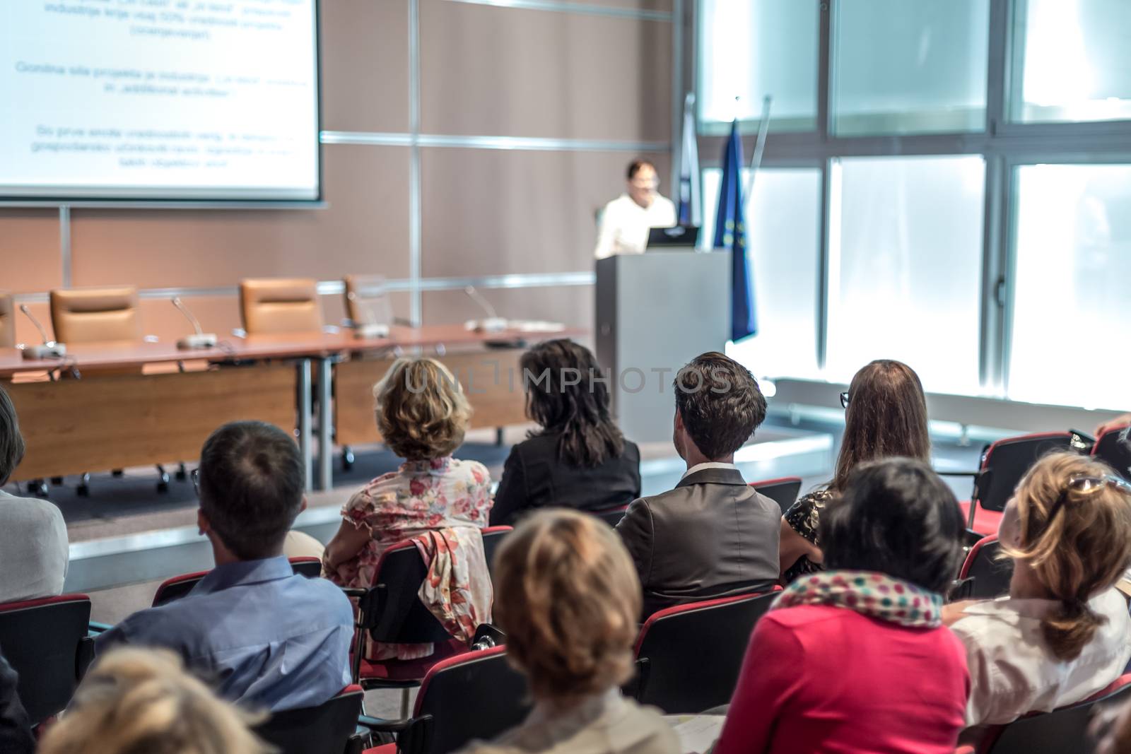 Female speaker giving presentation in lecture hall at university workshop. Audience in conference hall. Rear view of unrecognized participant in audience. Scientific conference event.