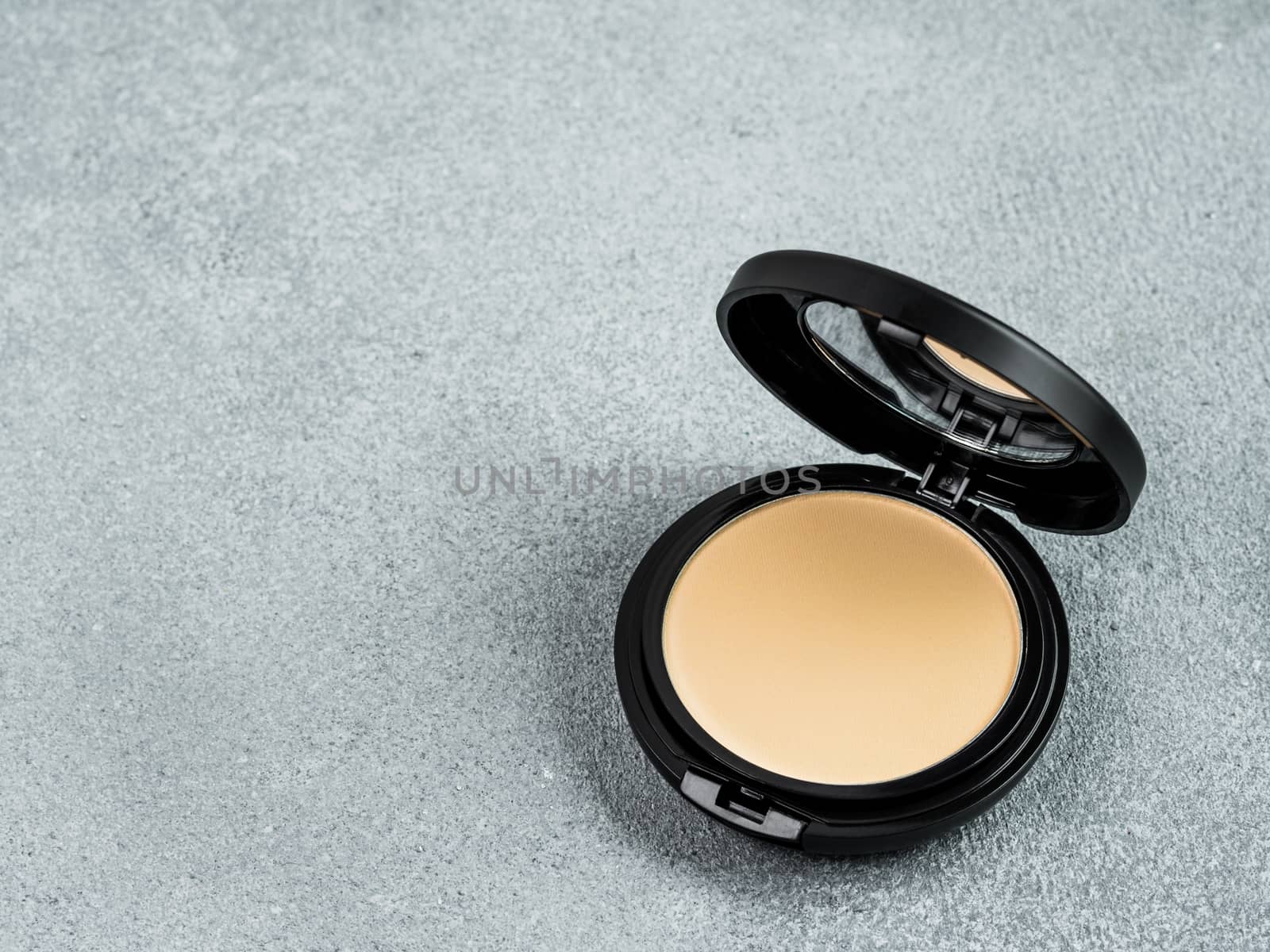 Compact powder on gray cement background. Female pressed powder in opened black plastic case with mirror, copy space for text or design