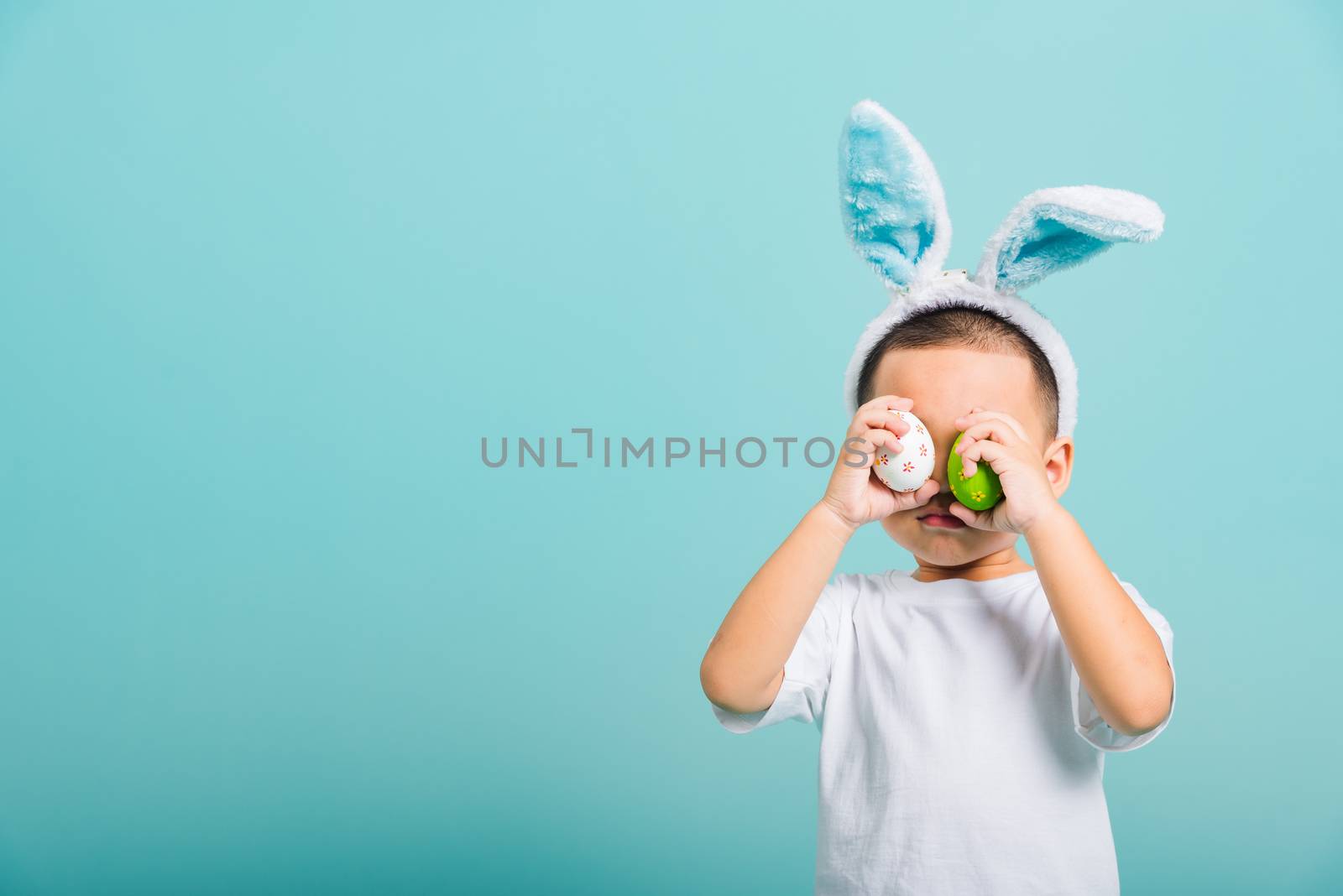 Asian cute little child boy smile beaming wearing bunny ears and a white T-shirt, standing to holds colored easter eggs instead of eyes on blue background with copy space for text