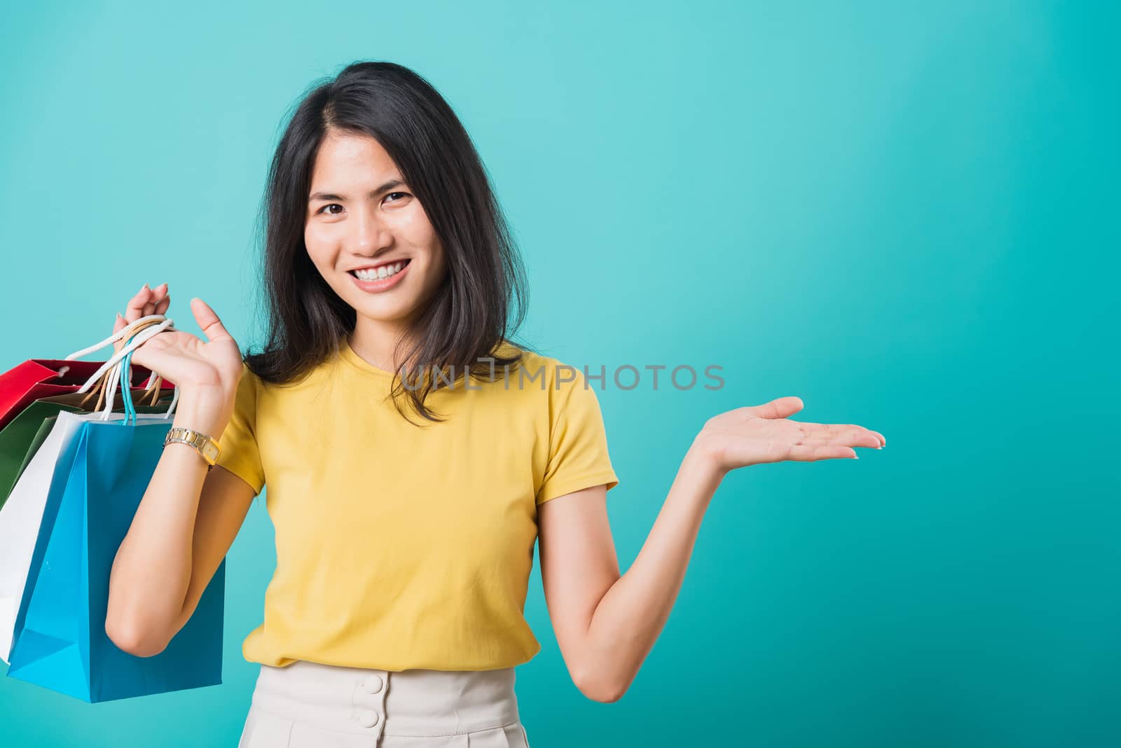 Portrait happy Asian beautiful young woman smile white teeth standing wear yellow t-shirt, She holding shopping bags show hand something empty and looking to camera, studio shot on blue background