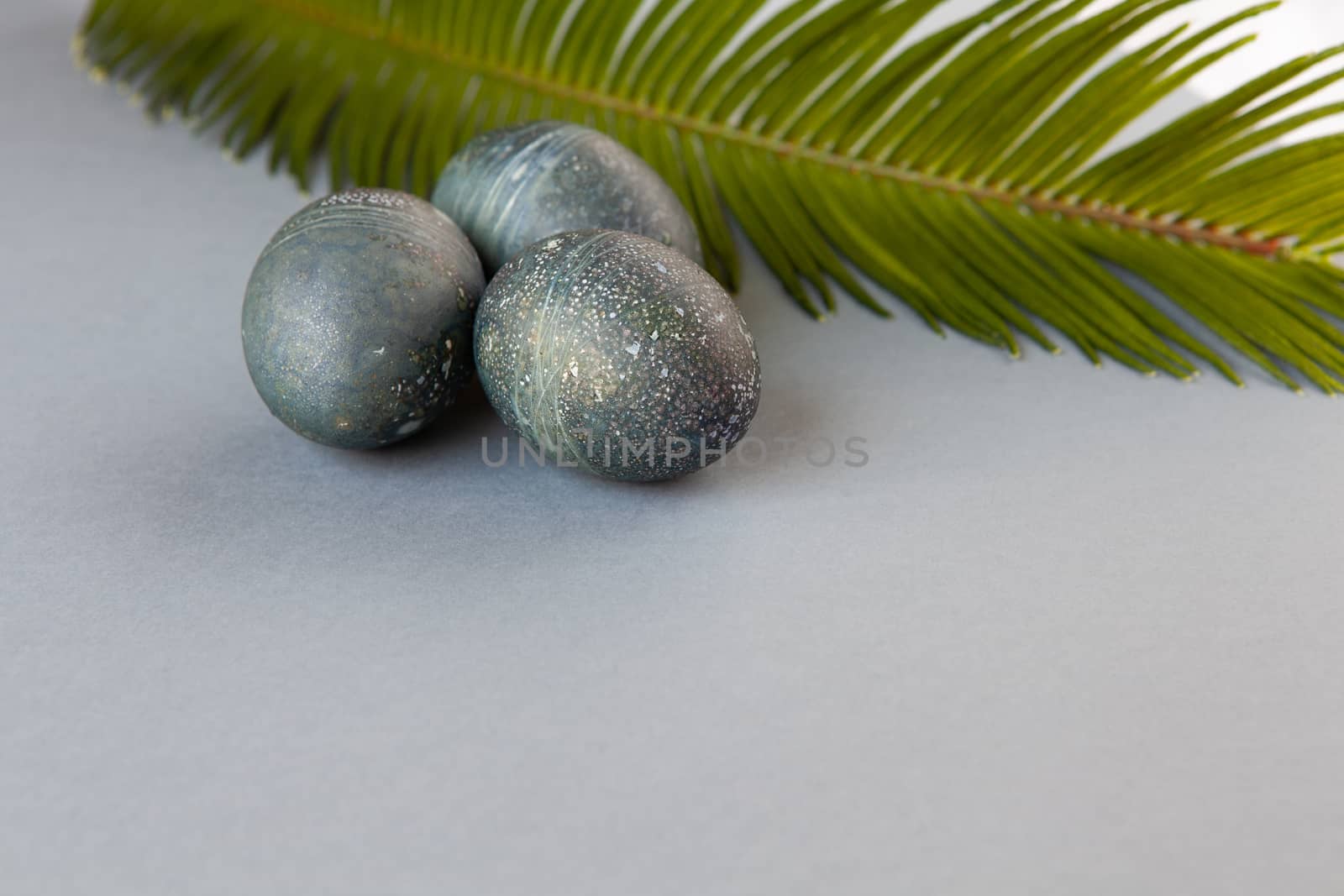 Creatively painted chicken Easter eggs with natural hibiscus dye, look like sea stones, date palm leaf on gray background, copy space. Happy creative Easter concept. Soft selective focus. Side view by ALLUNEED