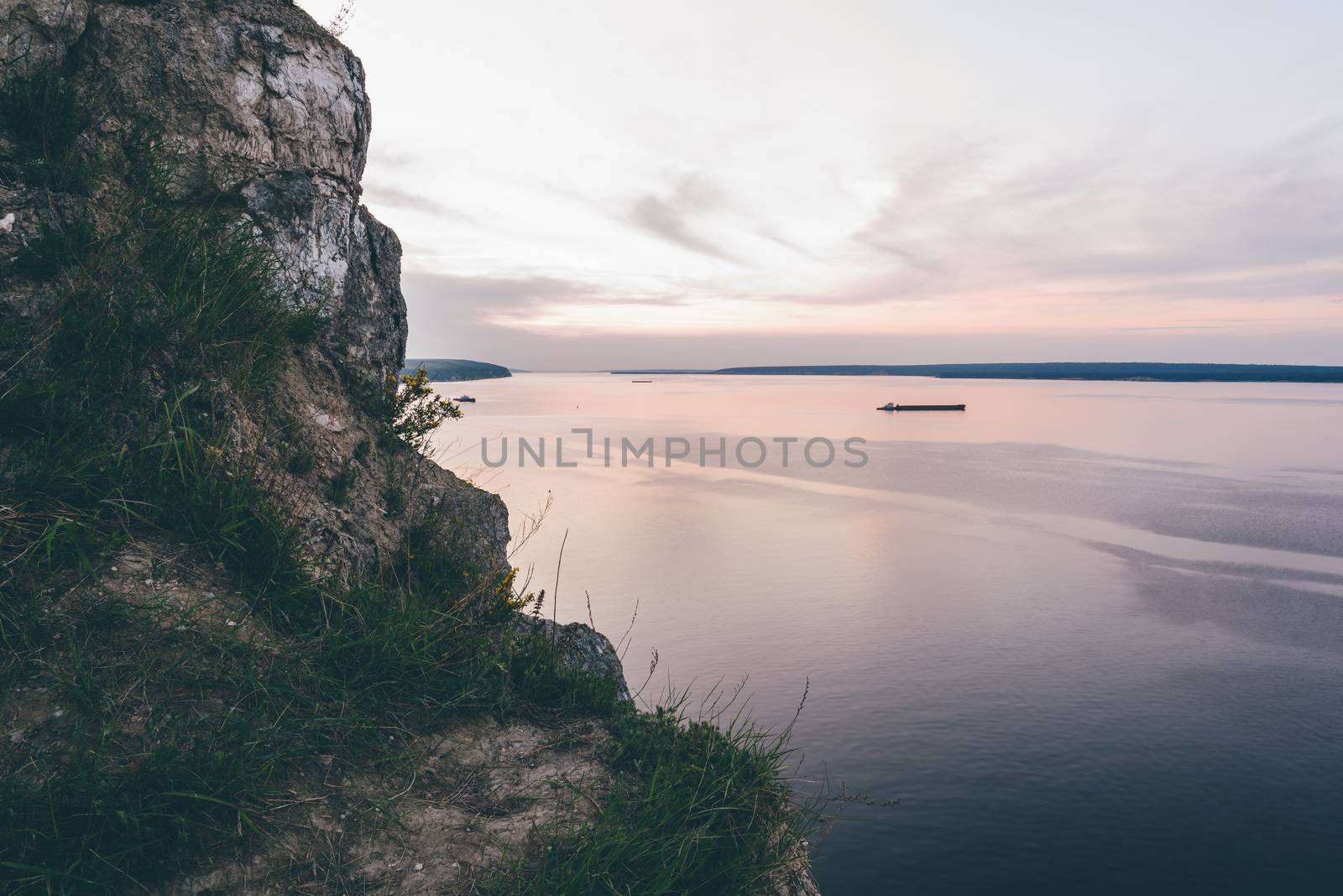 Cliff with grass and flowers with sunset on river on background