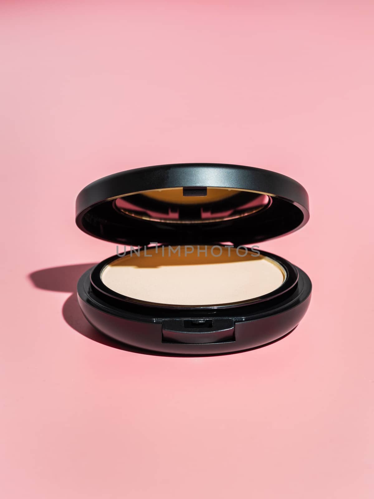 Compact powder on pink background. Female pressed powder in ajar opened black plastic case with mirror, copy space for text or design. Hard light. Vertical