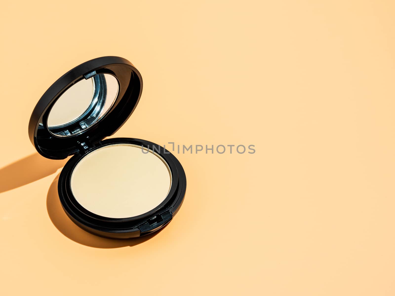 Compact powder on yellow or cream background. Female pressed powder in ajar opened black plastic case with mirror, copy space for text or design. Hard light