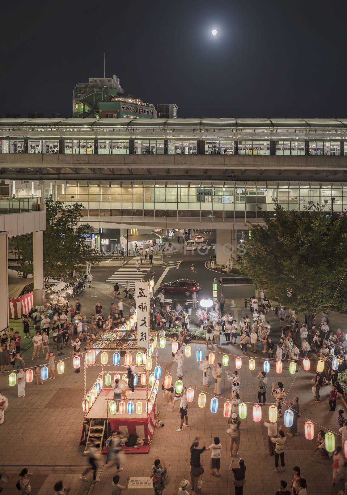 View of the square in front of the Nippori train station decorated for the Obon festival with a yagura tower illuminated with paper lanterns where a girl in traditional costume is playing taiko drum in the summer night of Arakawa district in Tokyo.