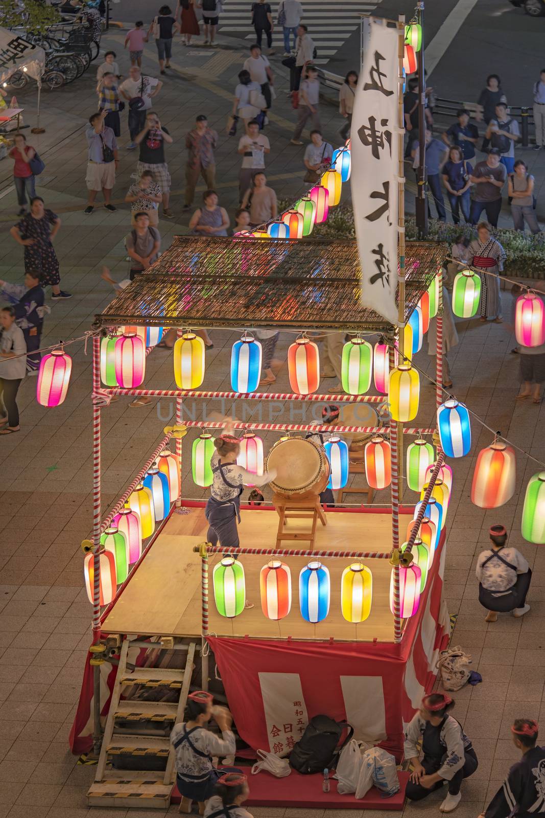 View of the square in front of the Nippori train station decorated for the Obon festival with a yagura tower illuminated with paper lanterns where a girl in traditional costume is playing taiko drum in the summer night of Arakawa district in Tokyo.