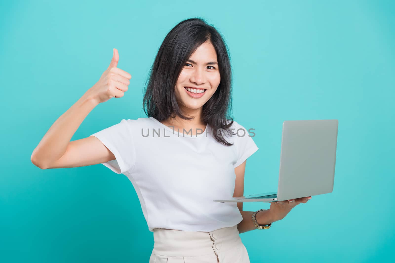 Asian beautiful young woman smile white teeth standing to holding laptop computer and showing thumb up while shoot photo in studio on blue background with copy space for text