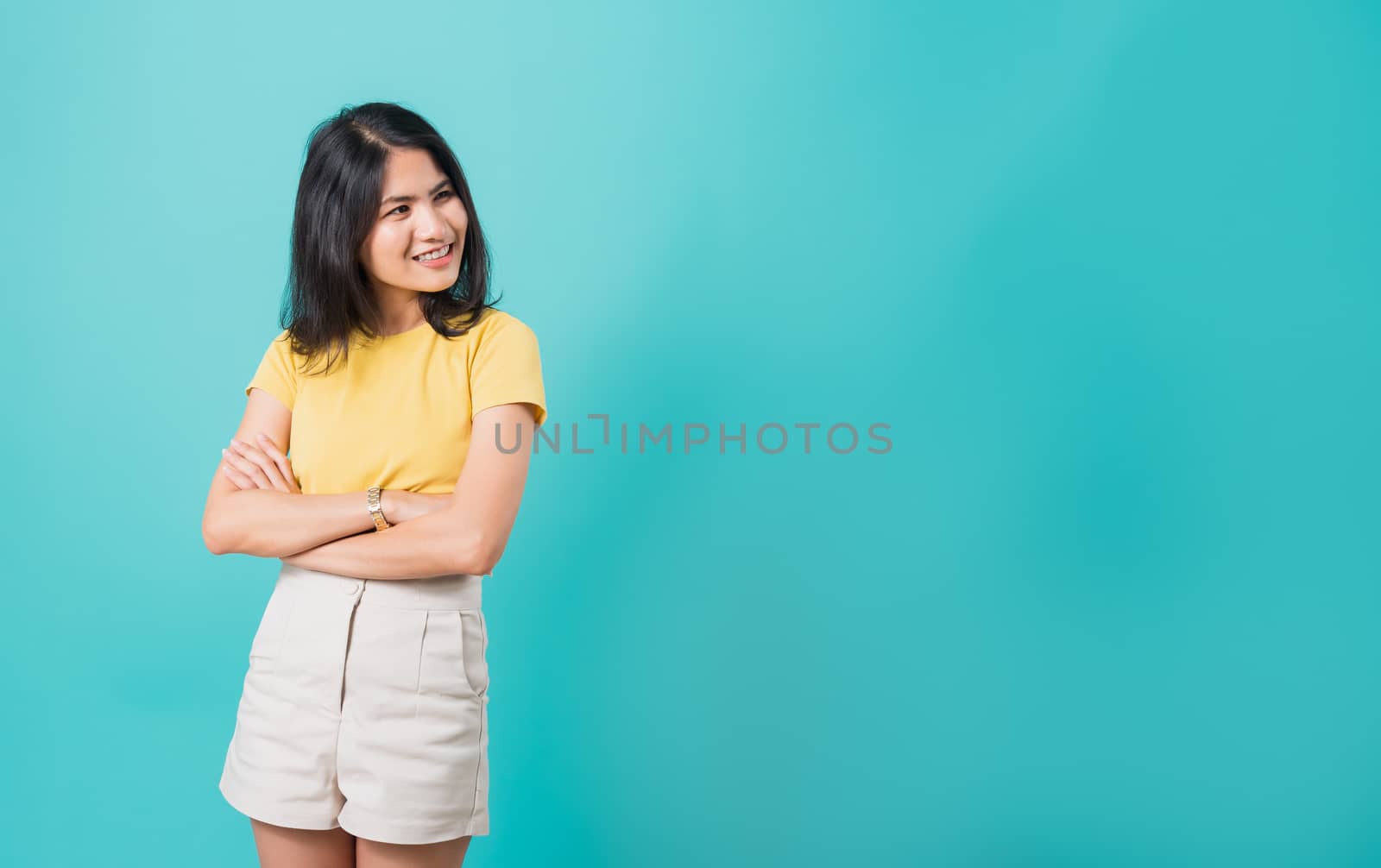 Portrait Asian beautiful happy young woman smiling wear yellow t-shirt standing cross arm with looking side away at copy space, shoot photo in a studio on a blue background