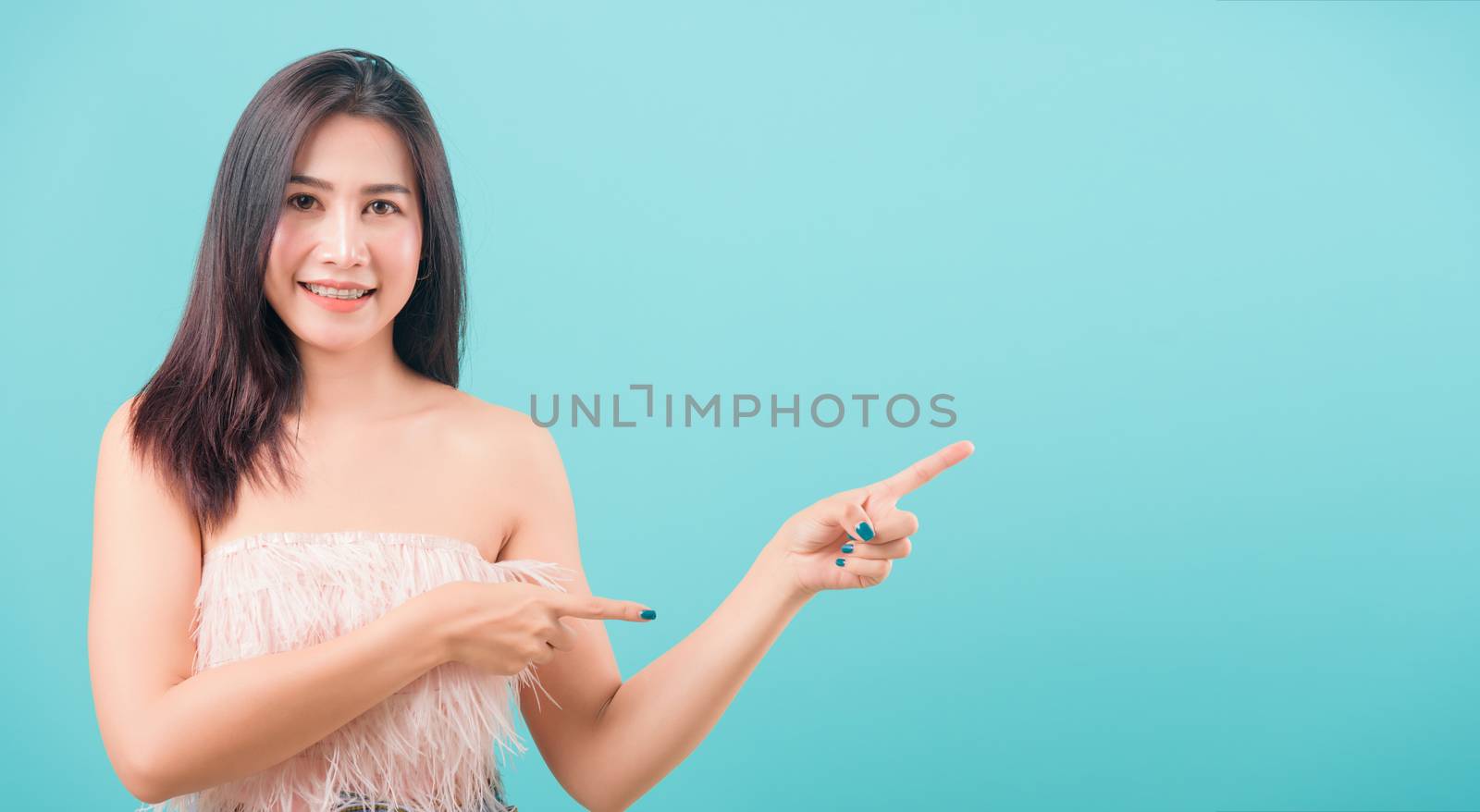 Asian happy portrait beautiful young woman standing her pointing finger outside and looking to camera isolated on blue background with copy space for text