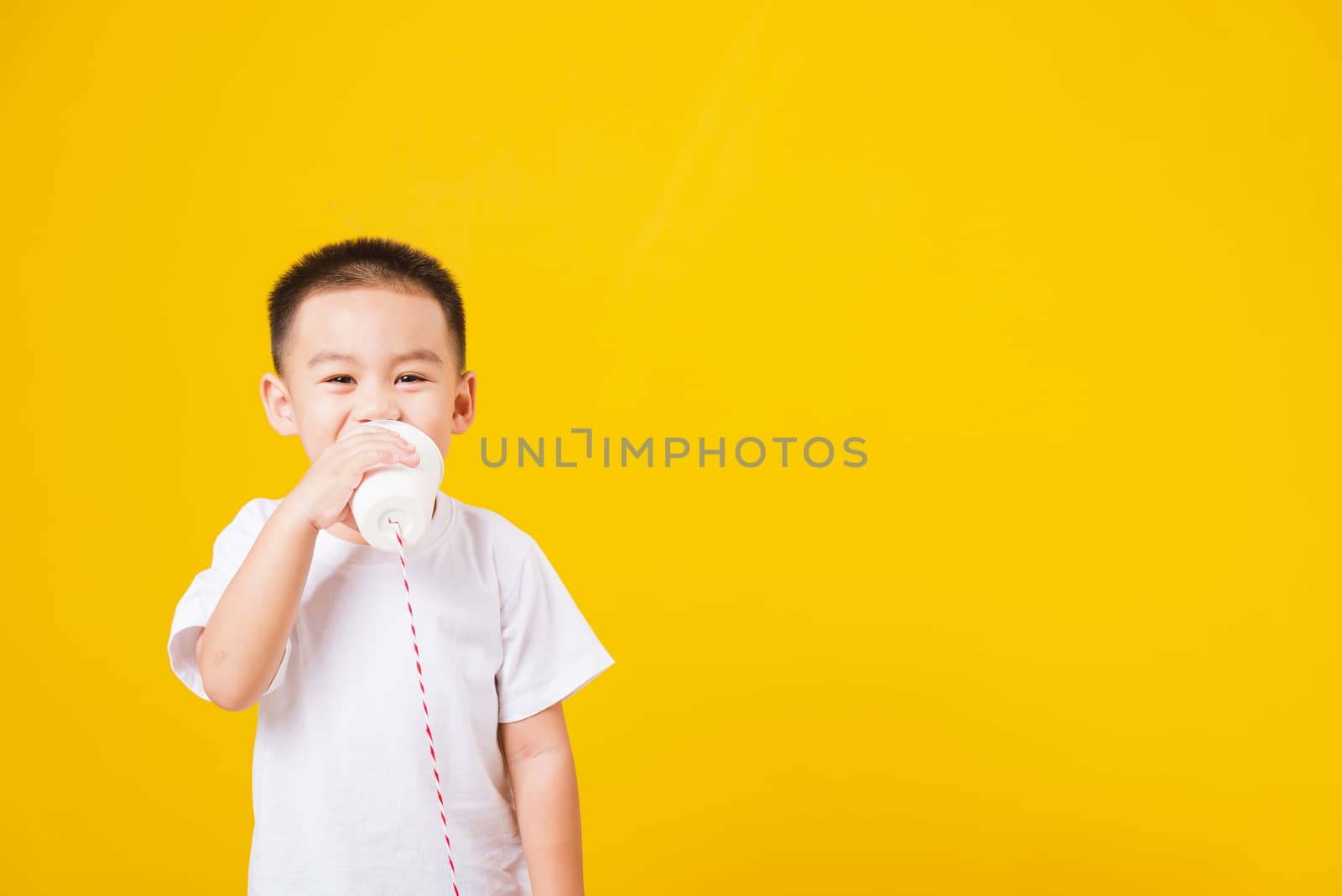 Portrait happy Asian cute little children boy smile standing so happy wearing white T-shirt playing paper can telephone, studio shot on yellow background with copy space