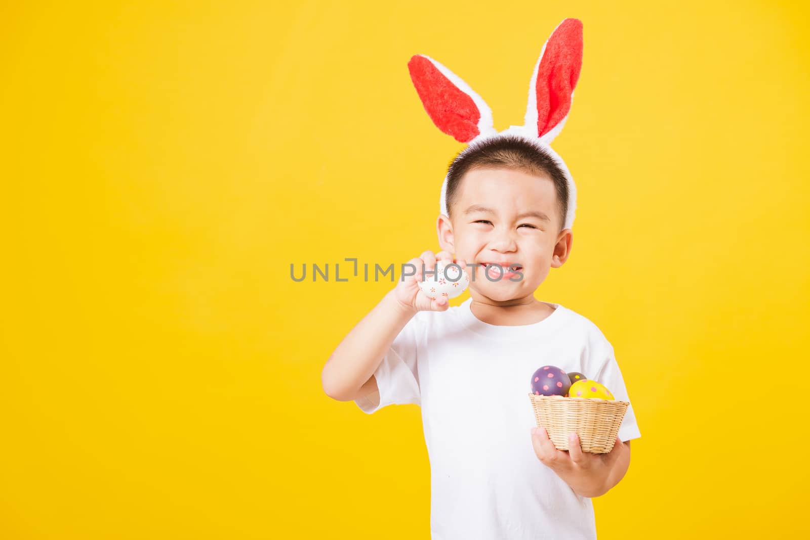 Portrait happy Asian cute little children boy smile standing so happy wearing white T-shirt and bunny ears in Easter festival day holding easter eggs, studio shot on yellow background with copy space