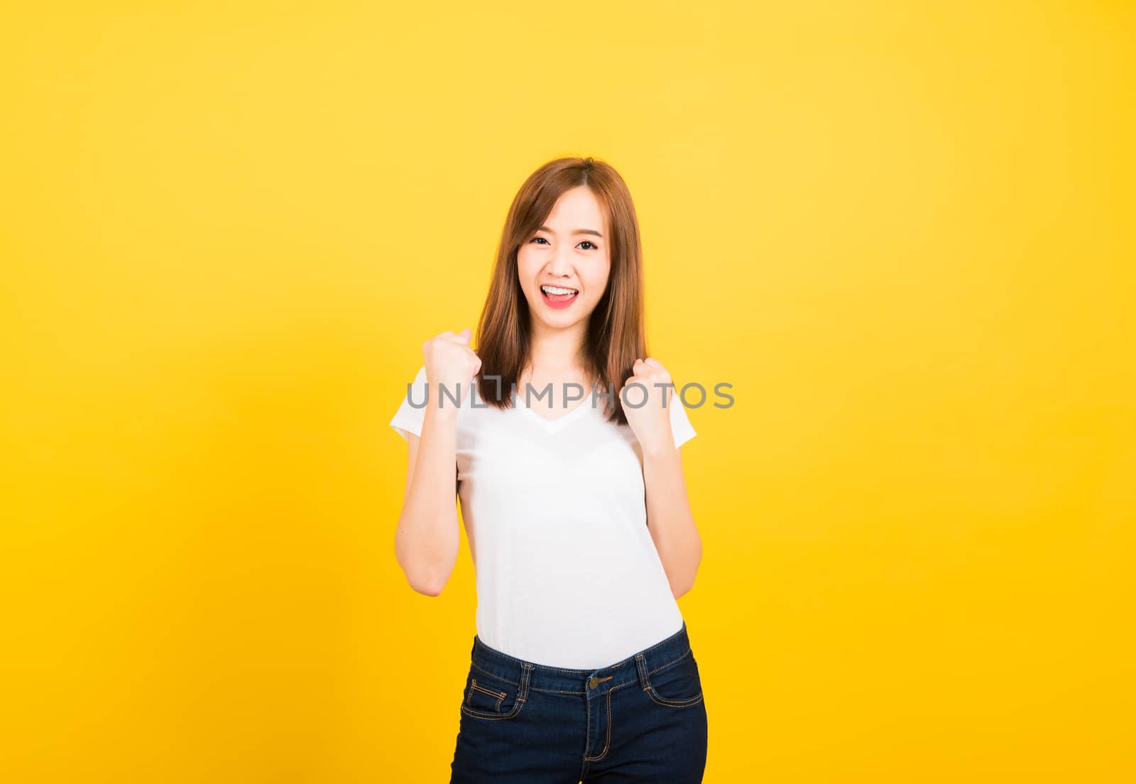 Asian happy portrait beautiful cute young woman teen standing wear t-shirt makes raised fists up celebrating her success looking to camera isolated, studio shot on yellow background with copy space