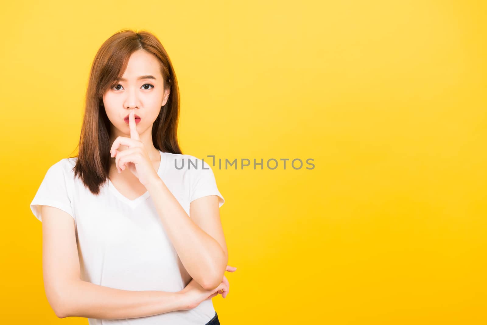 Asian happy portrait beautiful cute young woman teen smile standing wear t-shirt making finger on lips silent quiet gesture looking to camera isolated, studio shot on yellow background with copy space