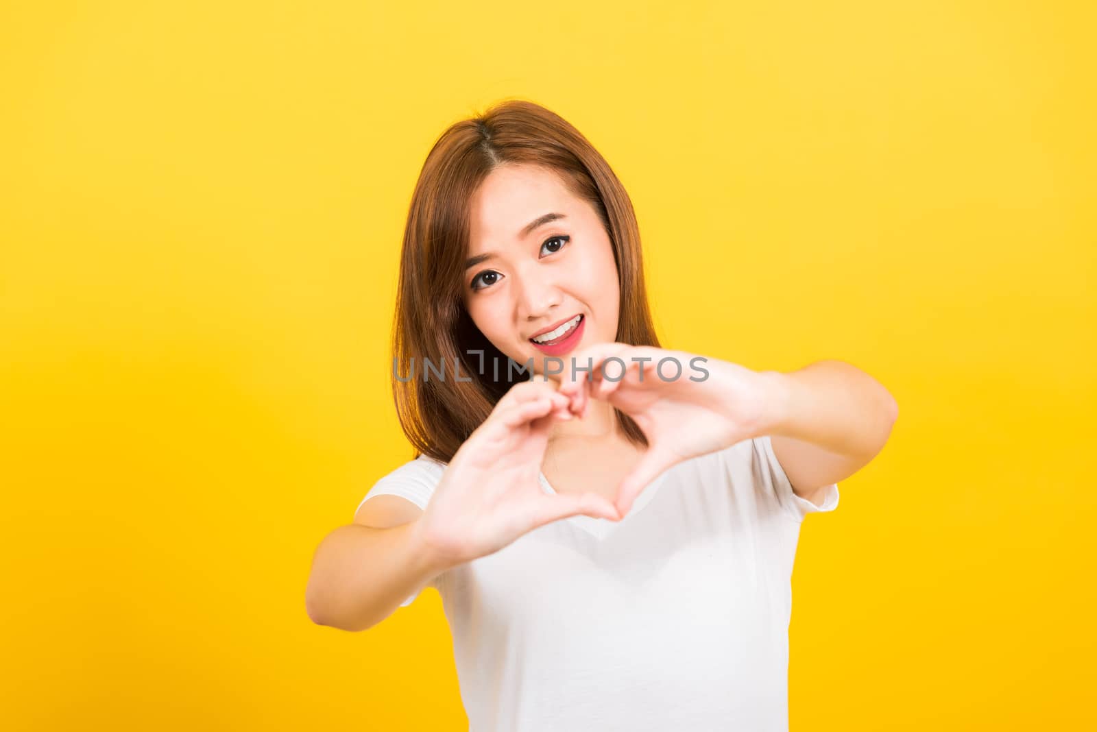 Asian happy portrait beautiful cute young woman teen smile standing make finger heart figure symbol shape sign with two hands looking to camera isolated, studio shot yellow background with copy space