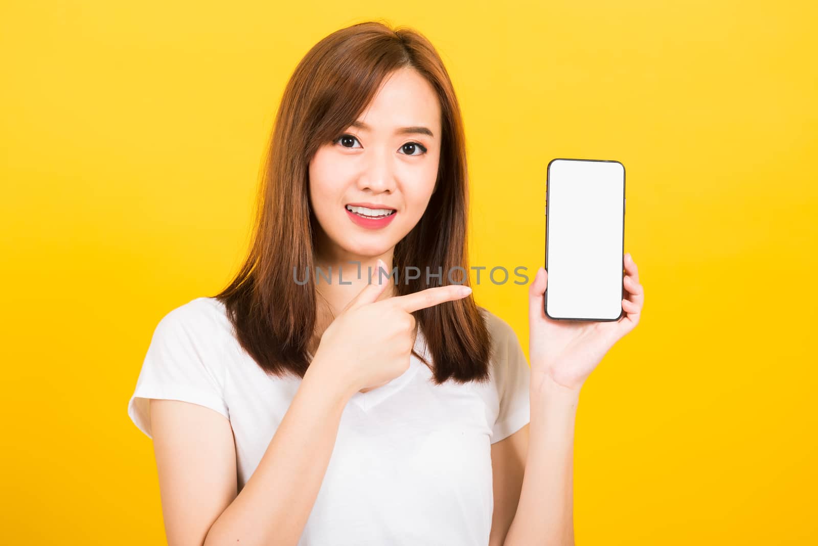 Asian happy portrait beautiful cute young woman smile standing wear t-shirt making finger pointing on smartphone blank screen looking to camera isolated, studio shot yellow background with copy space