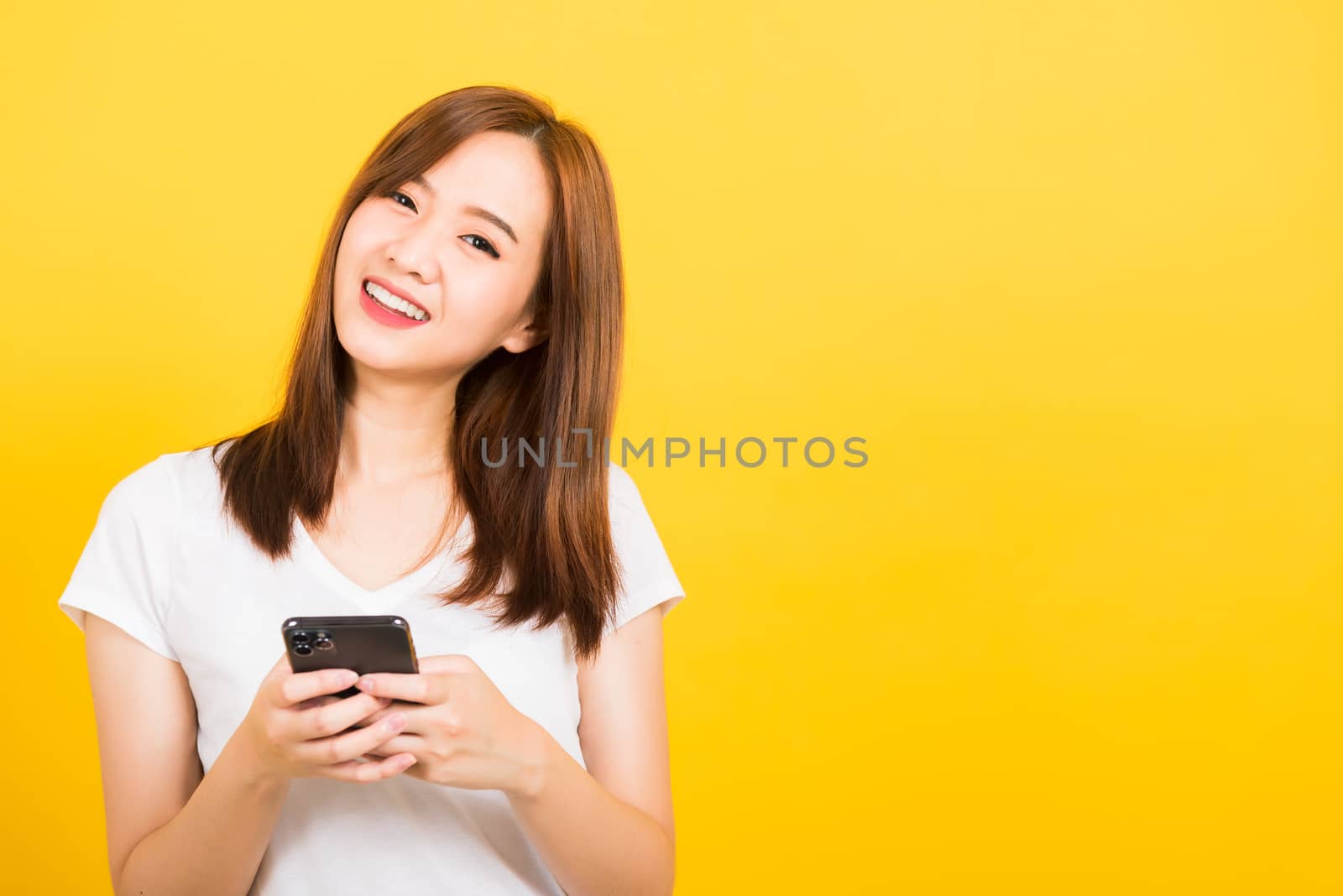 Asian happy portrait beautiful cute young woman teen smiling standing wear t-shirt using smart mobile phone looking to camera isolated, studio shot on yellow background with copy space