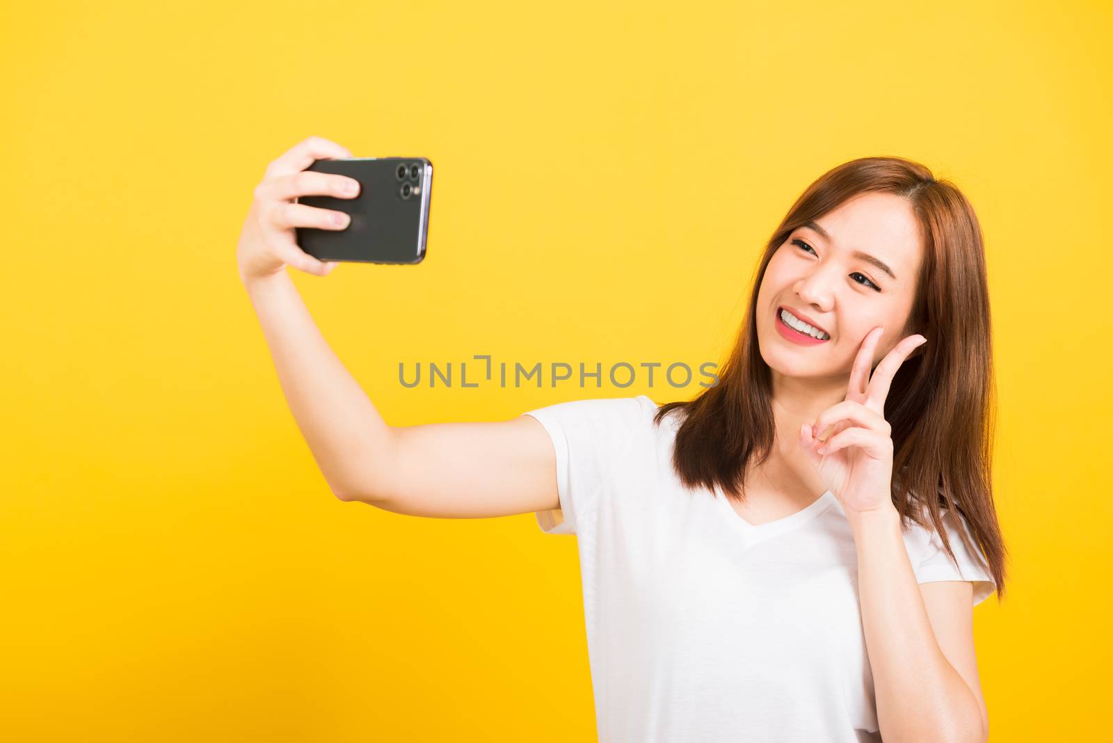 Asian happy portrait beautiful cute young woman teen smiling standing wear t-shirt making selfie photo, video call on smartphone looking camera isolated, studio shot yellow background with copy space