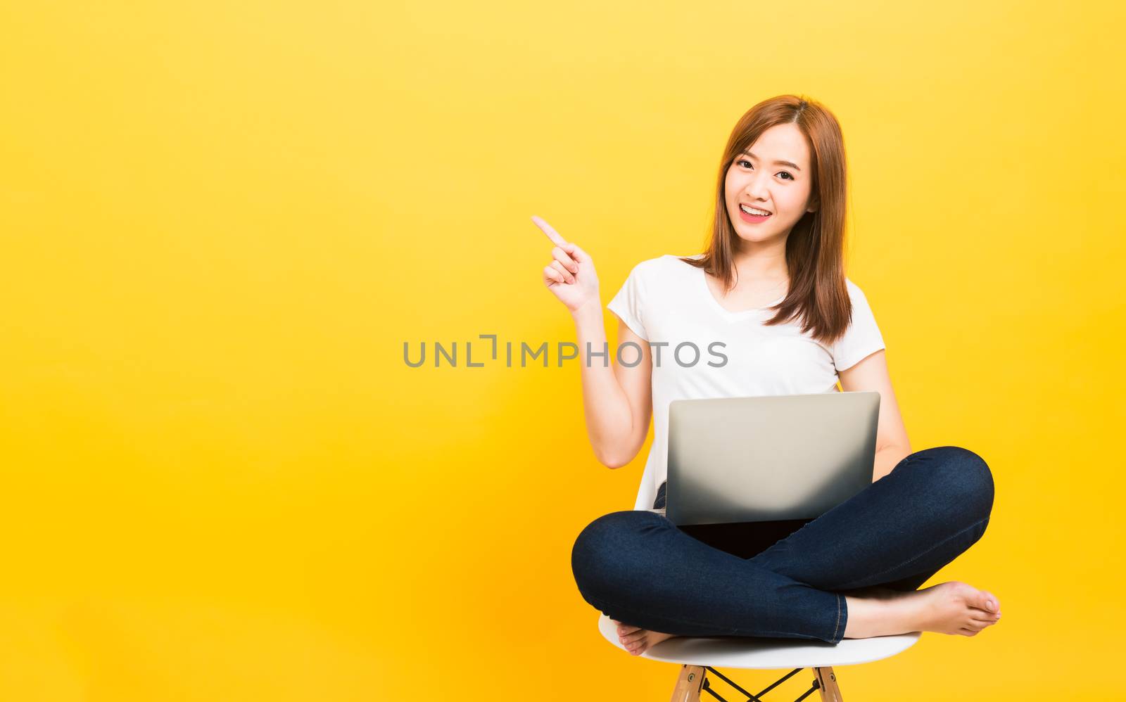 Asian happy portrait beautiful cute young woman teen smiling sitting crossed legs on chair with laptop computer point finger side looking camera isolated, studio shot yellow background with copy space