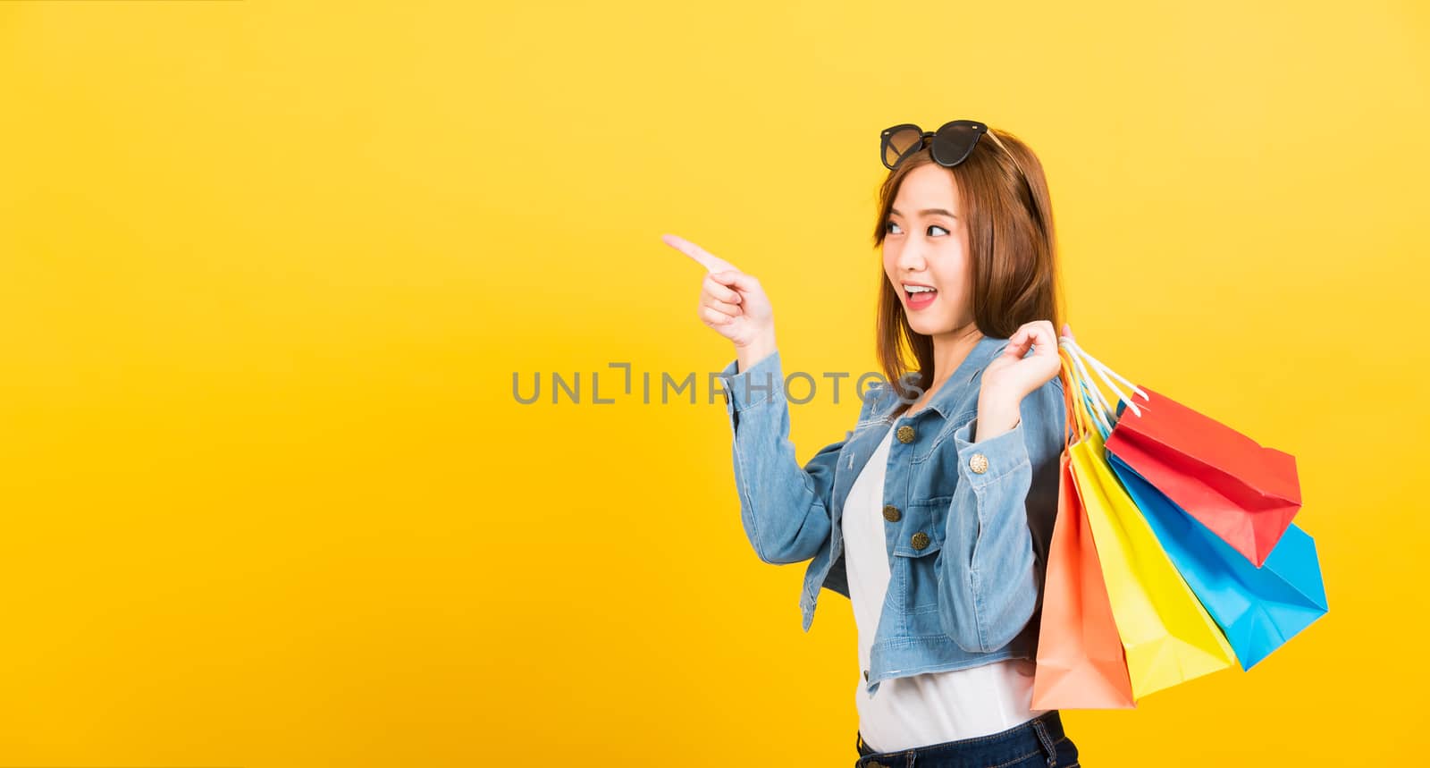 Asian happy portrait beautiful cute young woman teen smiling standing with sunglasses excited holding shopping bags multi color pointing to side isolated, studio shot yellow background with copy space