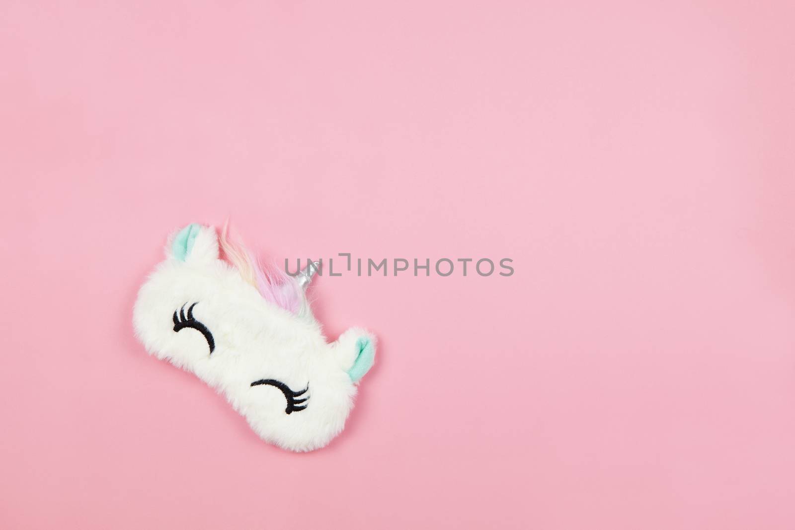 White fluffy fur sleep mask unicorn with closed eyes and small ears on pastel pink paper background, copy space. Top view, flat lay. Concept of vivid dreams. Accessories for girls and young women.