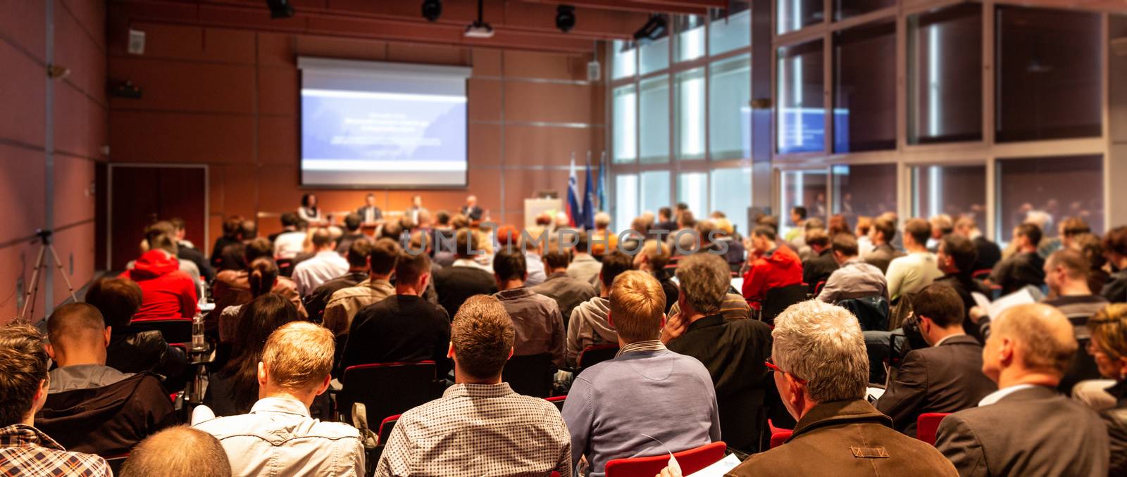 Business and entrepreneurship symposium. Speaker giving a talk at business meeting. Audience in conference hall. Rear view of unrecognized participant in audience.