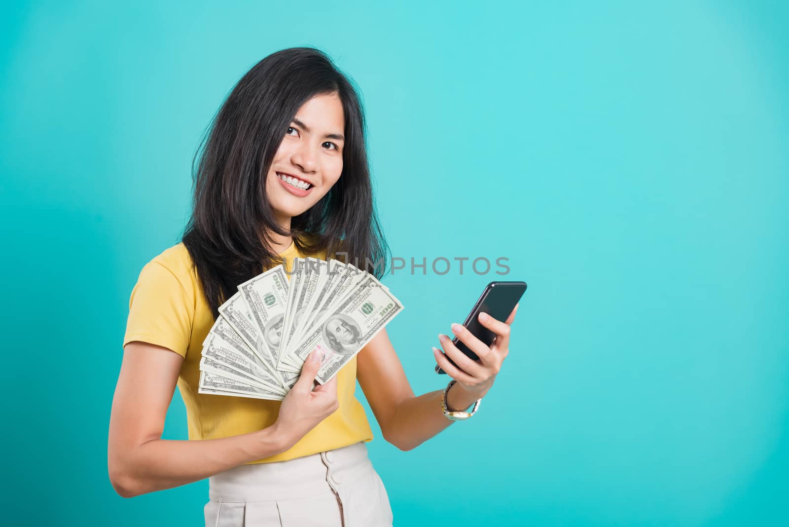 Asian happy portrait beautiful young woman standing wear t-shirt smile holding money fan banknotes 100 dollar bills using mobile phone and looking to camera on blue background with copy space for text